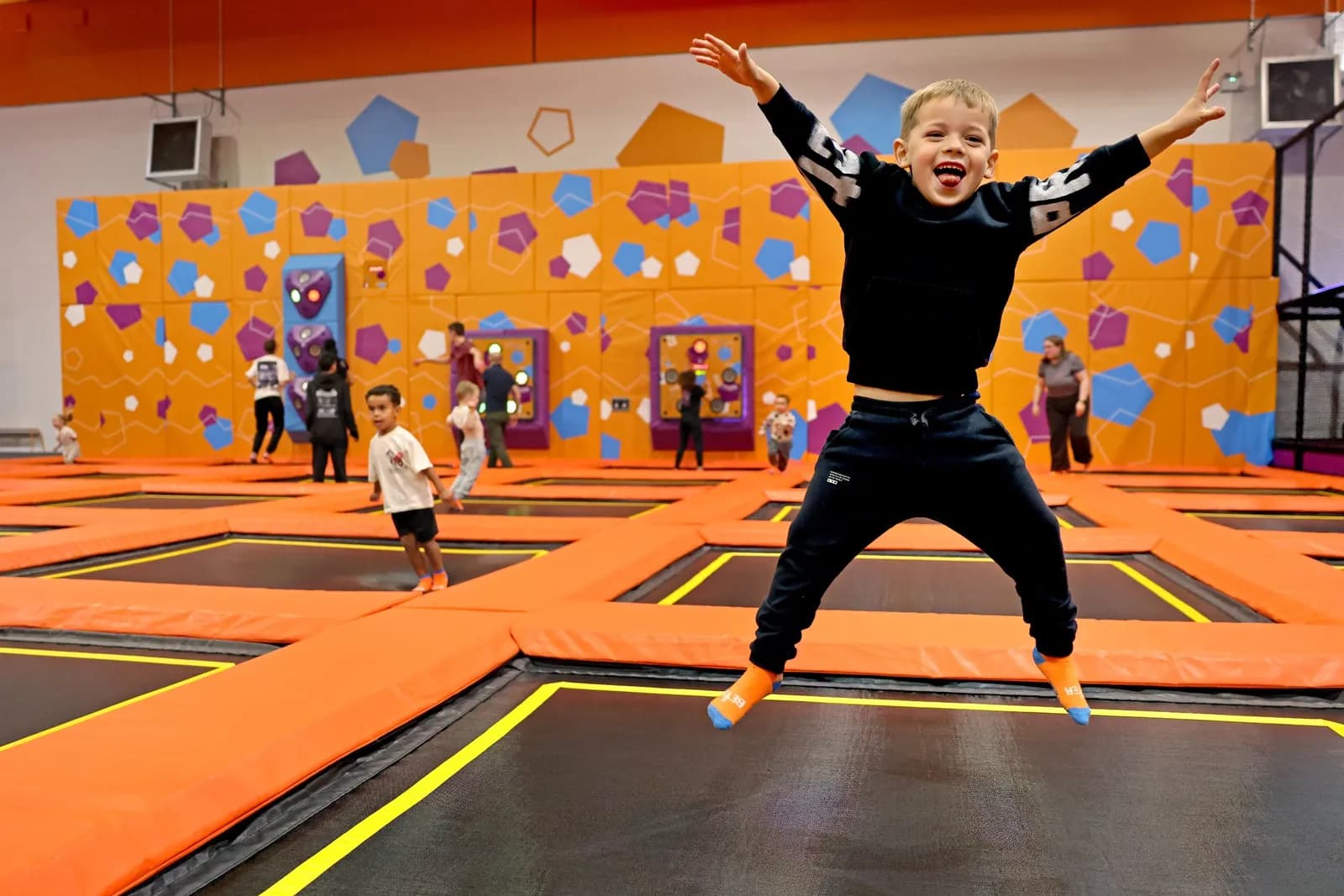 A young boy in a black long-sleeved shirt and pants leaps high in the air on a trampoline, with his arms outstretched and a wide grin on his face, while other children and adults are visible on other trampolines in the background.