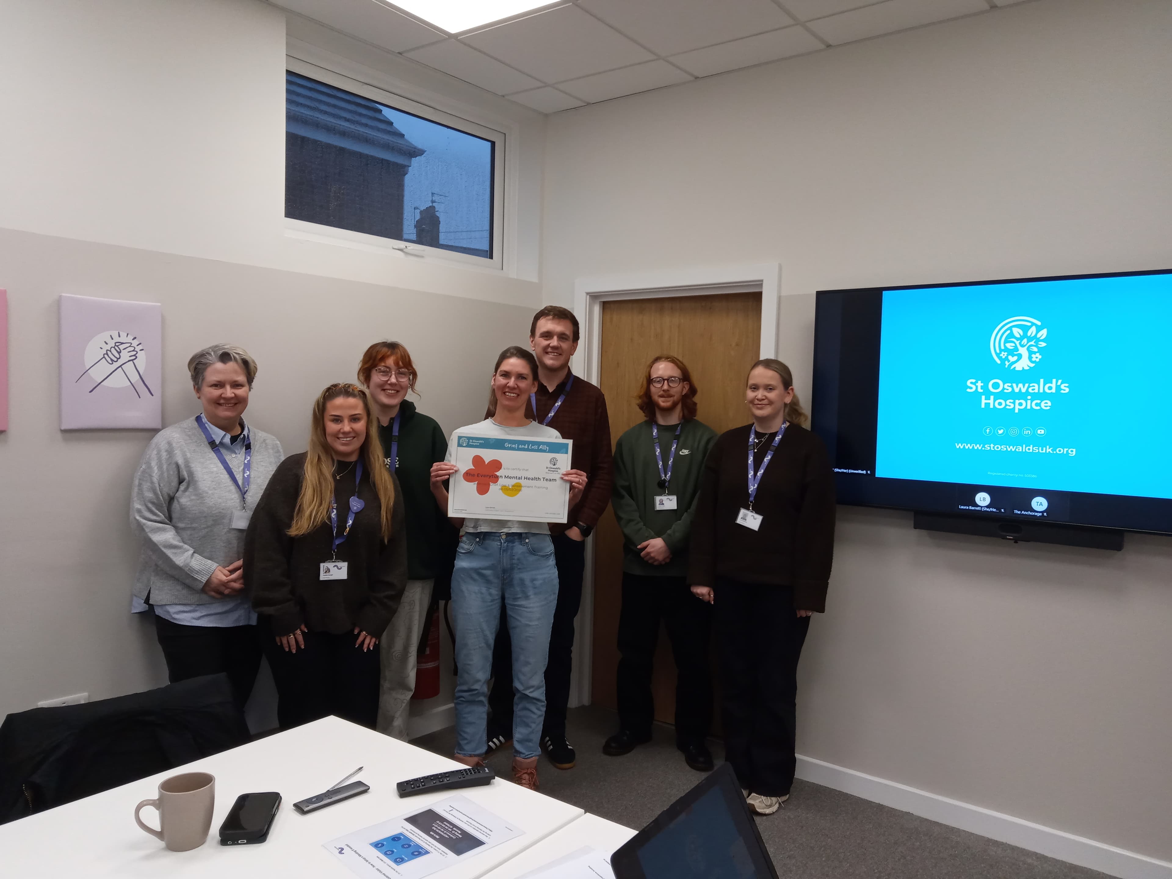 A group of seven people gathered in an office, with one woman holding a certificate while standing in front of a monitor displaying "St Oswald's Hospice."
