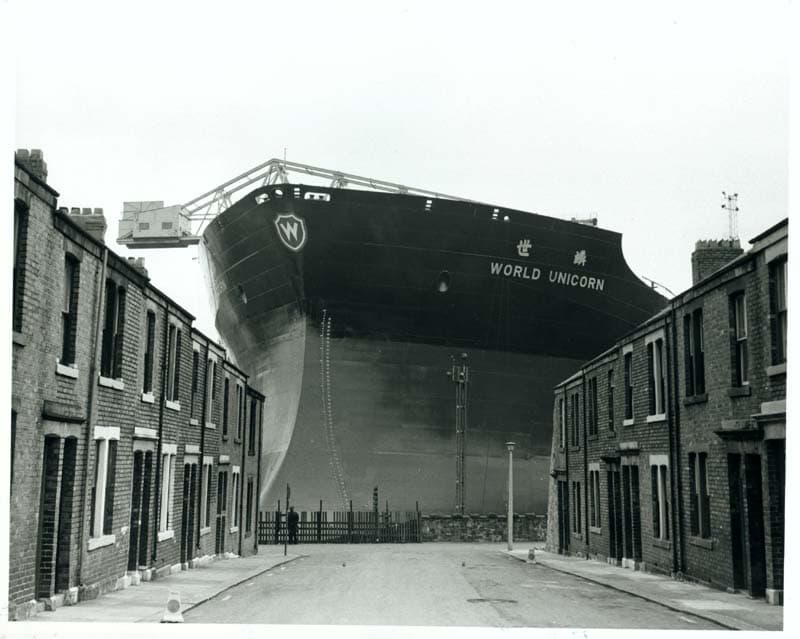 A towering ship named 'World Unicorn' stands prominently behind a row of brick houses, its massive hull dominating the skyline.