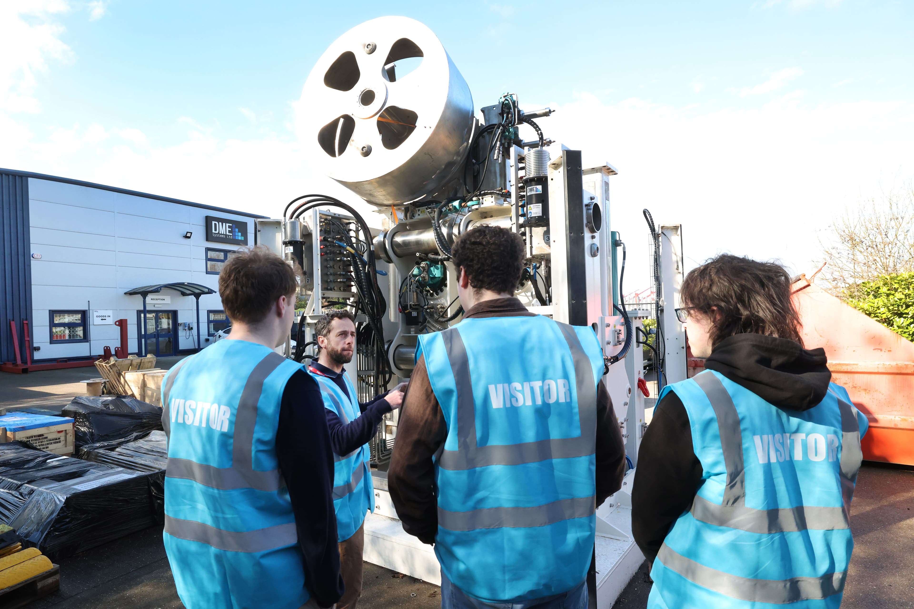 A man in a blue vest with "VISITOR" written on the back discusses a large piece of industrial machinery with three other people, also wearing blue visitor vests, in an outdoor setting next to a building.