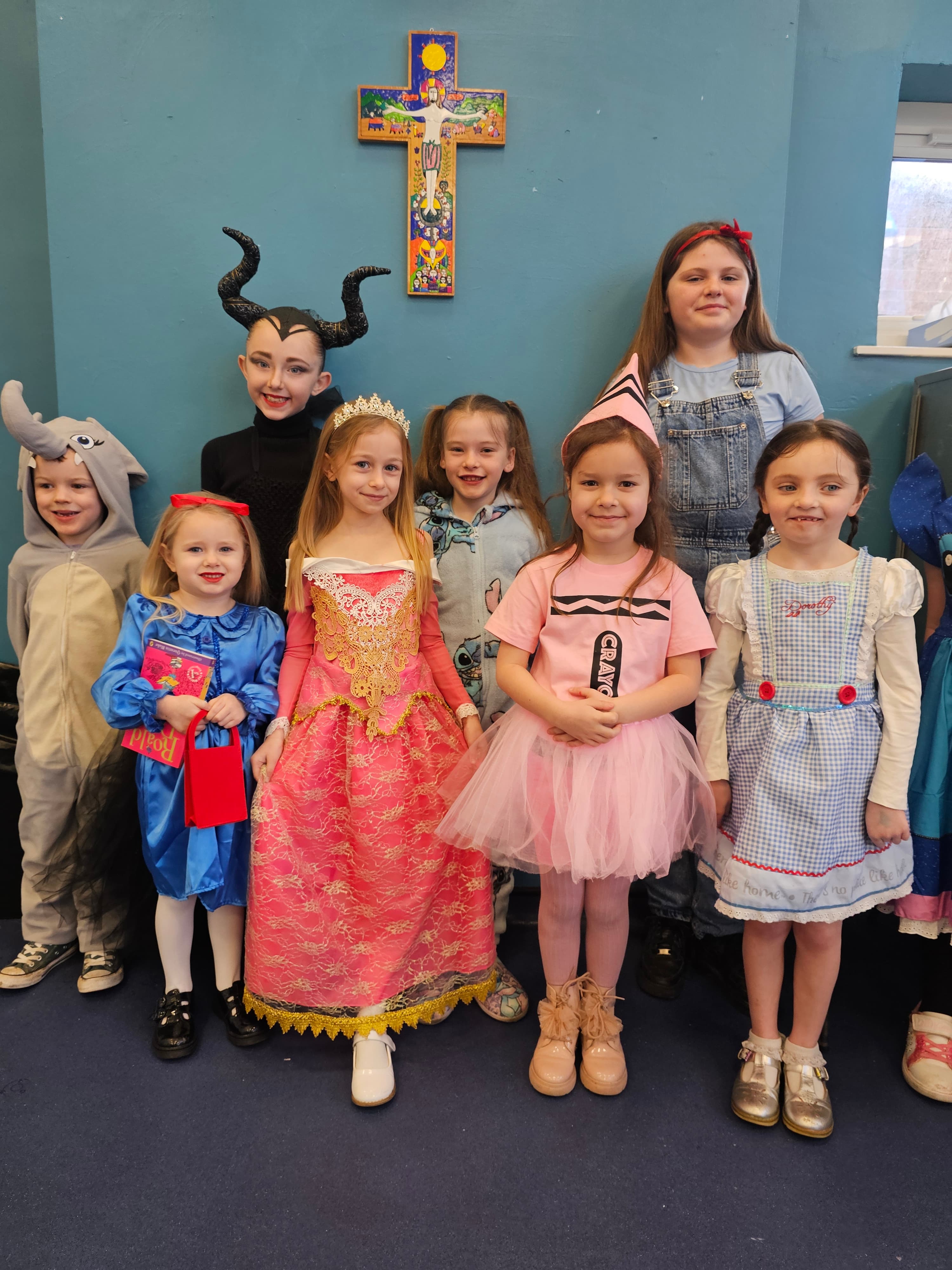 A group of children in various costumes including a princess, a Maleficent-like figure, Dorothy from the Wizard of Oz, a crayon, an elephant, and a little girl in a blue dress with a book, posing indoors with a cross on the wall.
