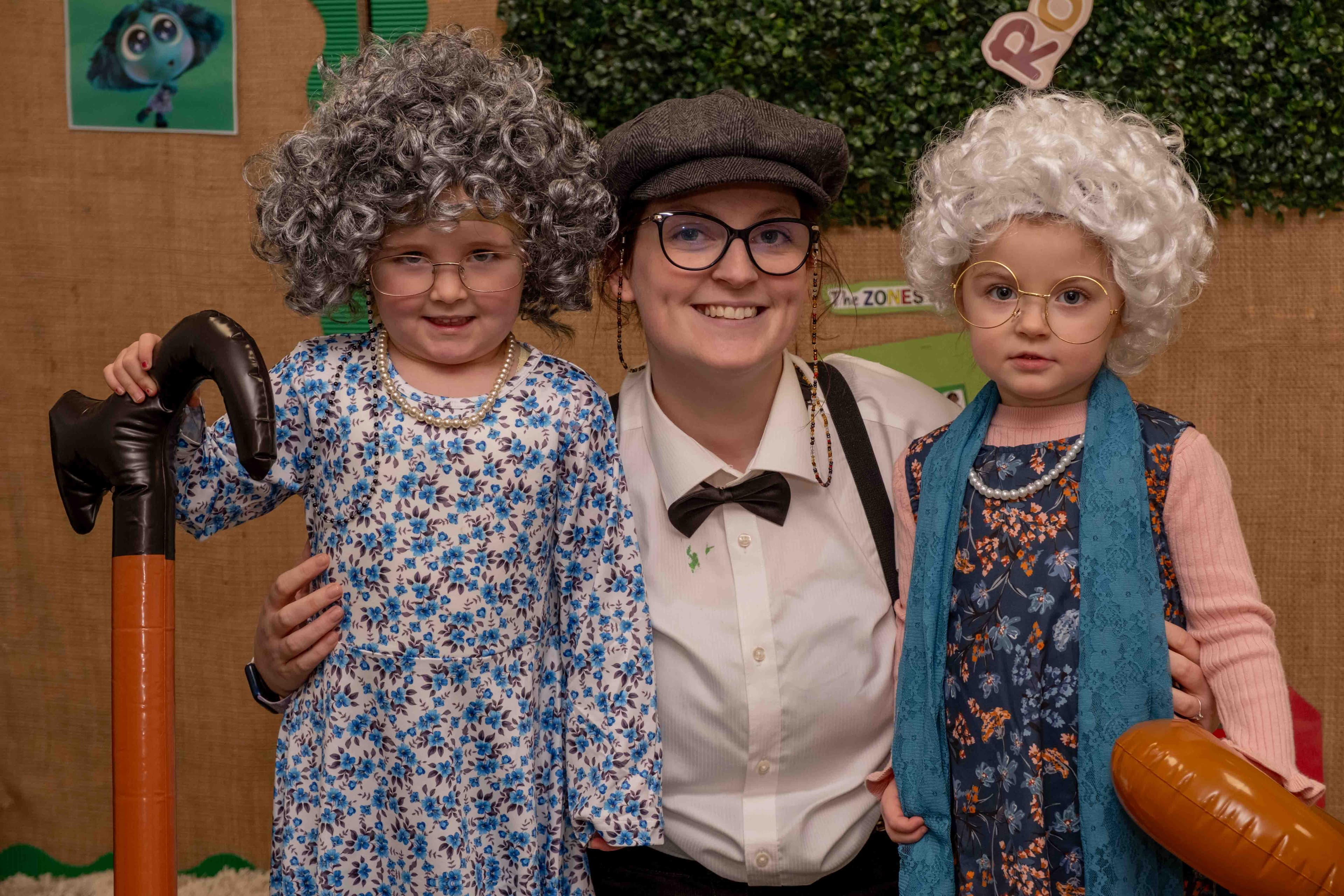 A smiling young person in a flat cap and bow tie stands between two small children dressed as old women with grey curly wigs, glasses, and pearl necklaces, one holding an inflatable cane.
