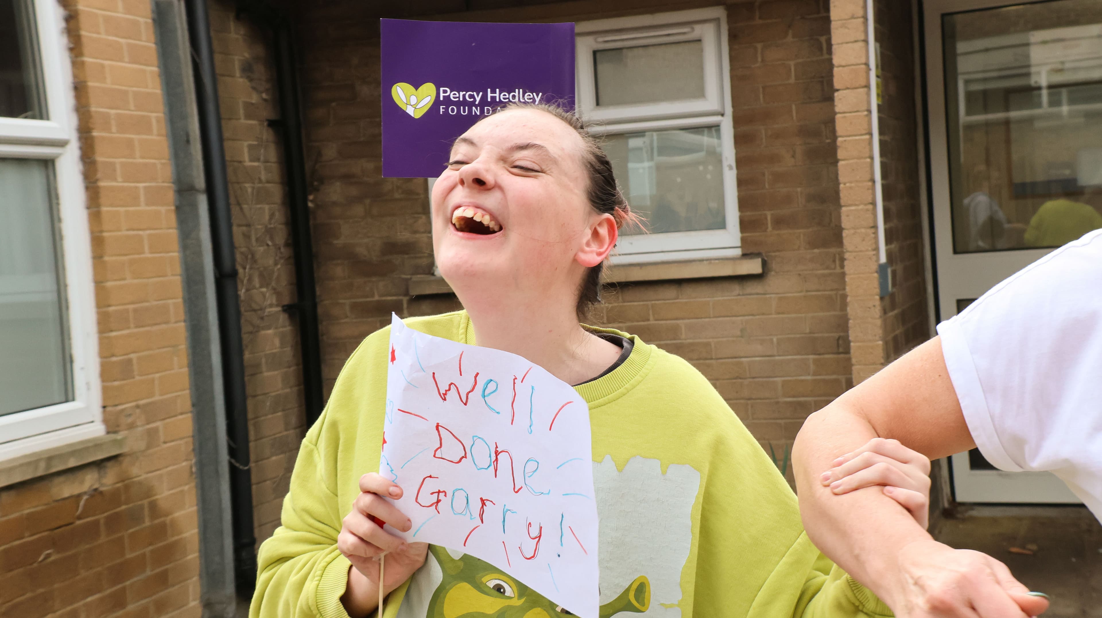 A young person with a wide open, joyful laugh holds a sign that says 'Well Done Garry!' with a purple Percy Hedley Foundation sign in the background.