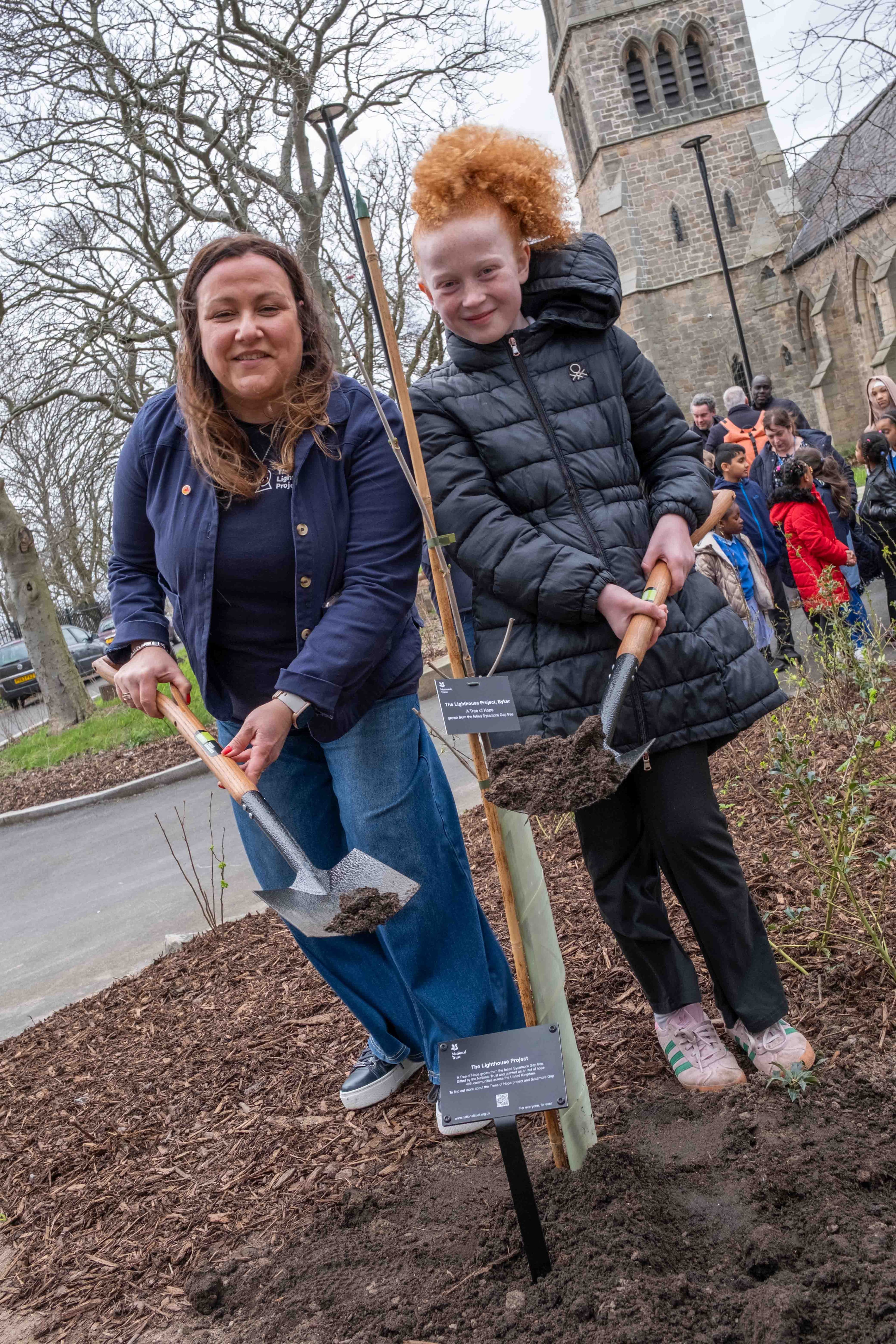 A woman and a young girl with red hair, both holding shovels with dirt on them, stand next to a newly planted tree with a small sign.