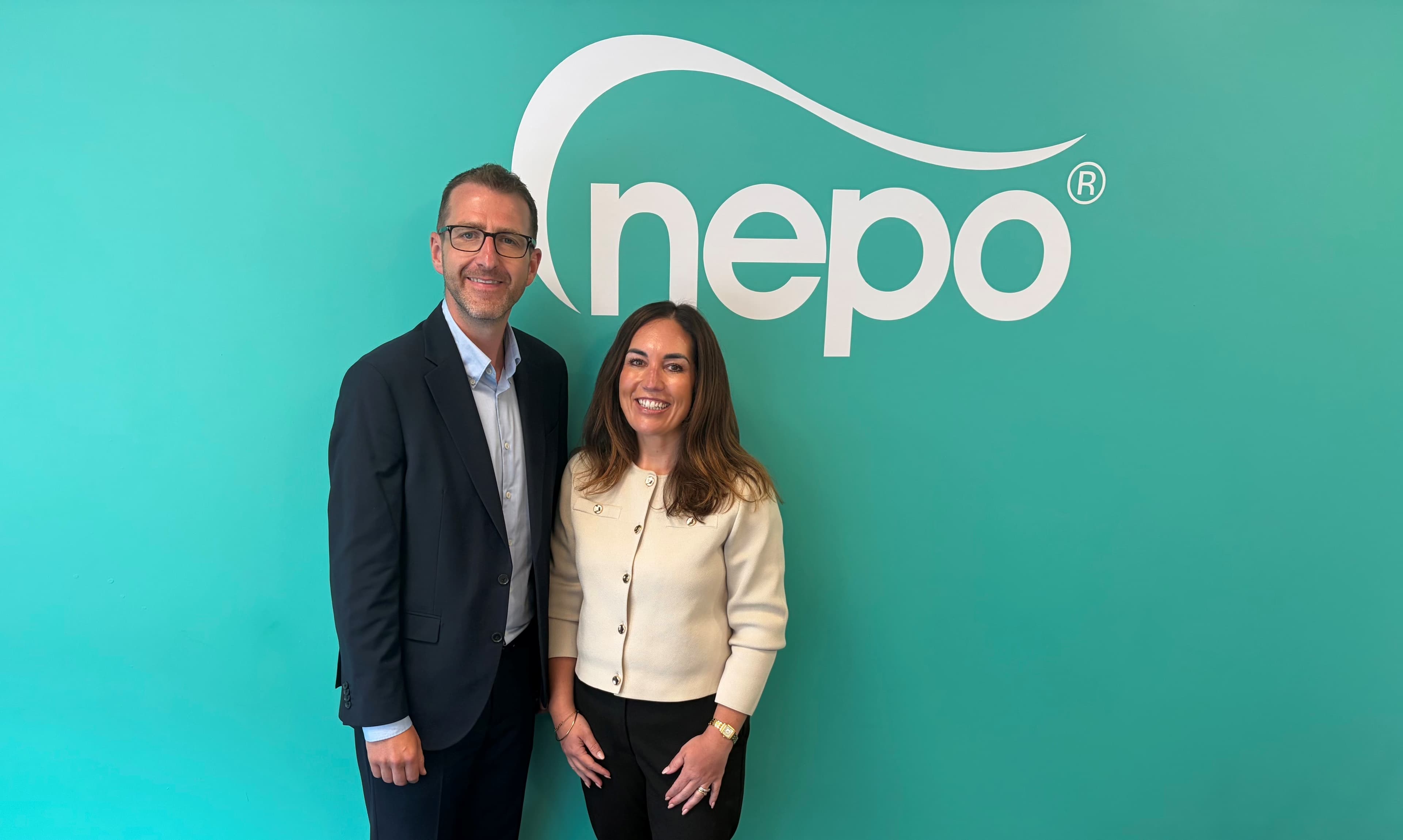 A man in a suit and a woman in a light-colored cardigan stand smiling in front of a teal wall with a white 'nepo' logo.