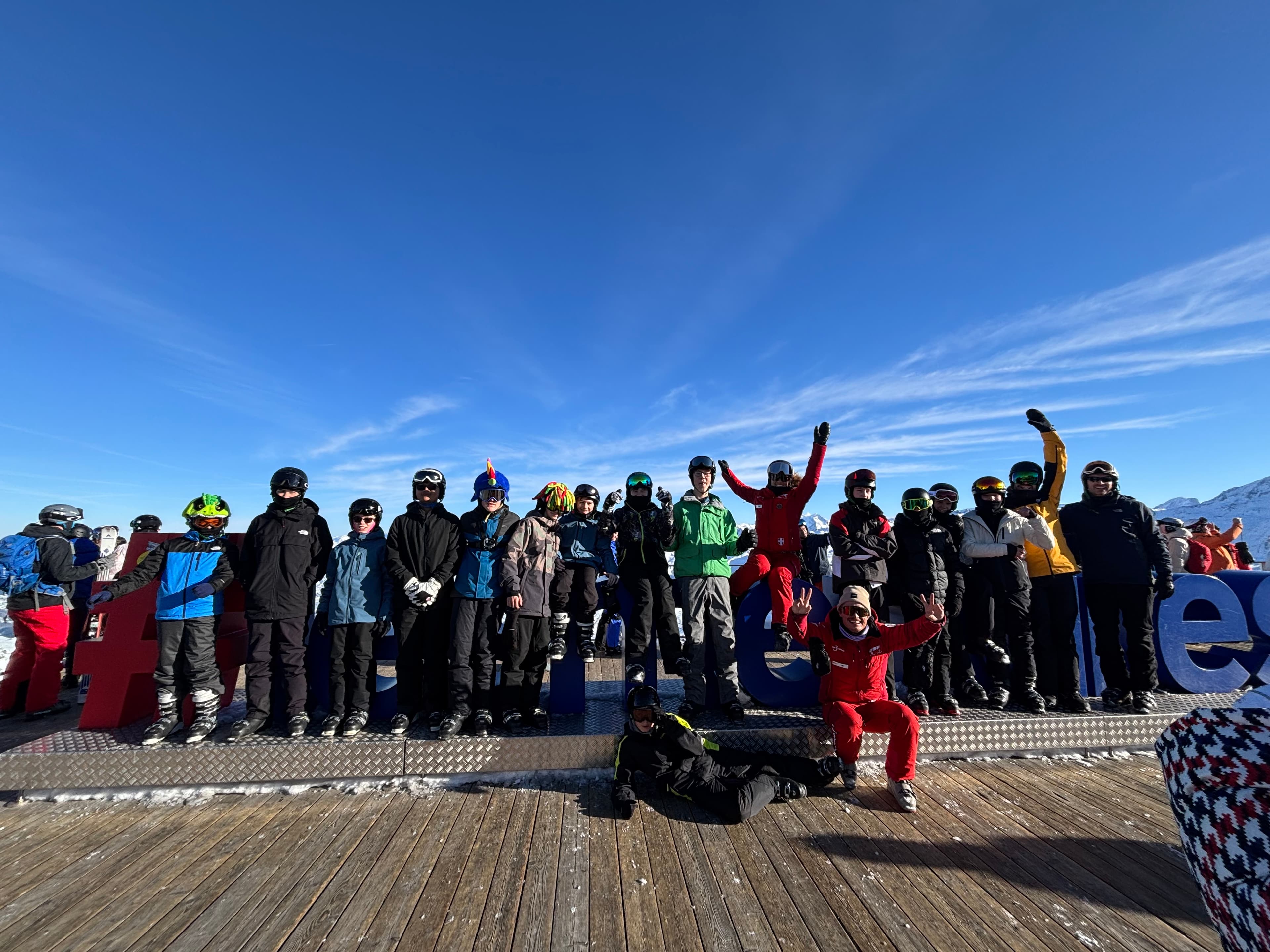 A group of happy skiers and snowboarders pose for a photo on a wooden deck with a bright blue sky behind them.