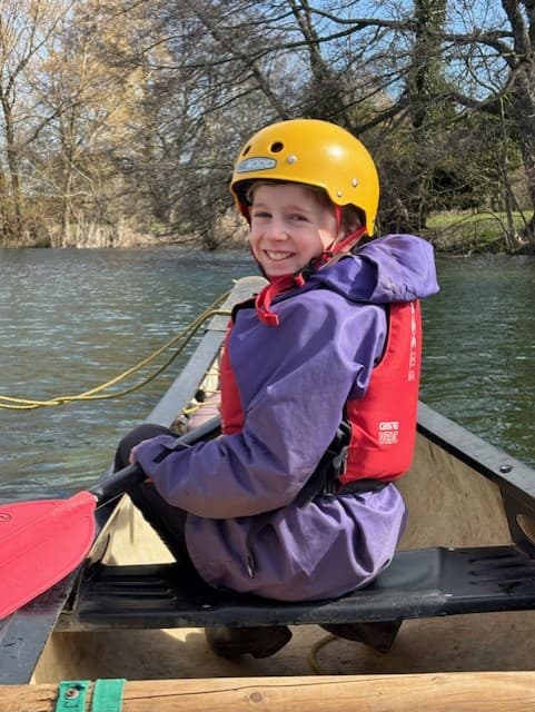 A young boy wearing a yellow helmet, purple jacket, and red life vest smiles at the camera from inside a canoe on a sunny day.