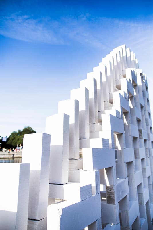 A modern architectural structure made of white blocks is shown against a clear blue sky, with some greenery and water visible in the background.