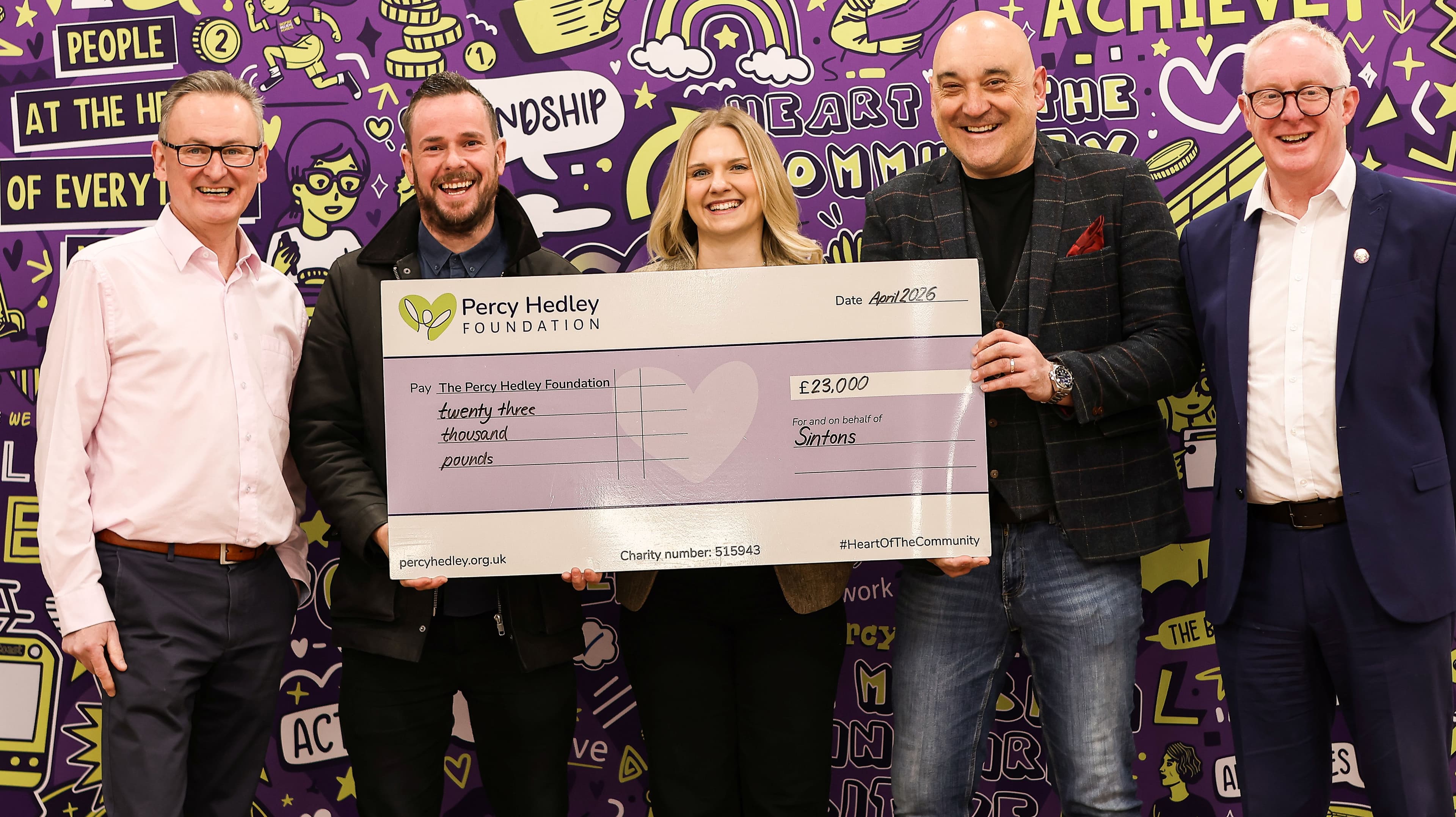 Five adults, three men and one woman, smile while holding a large check for £23,000 made out to The Percy Hedley Foundation.