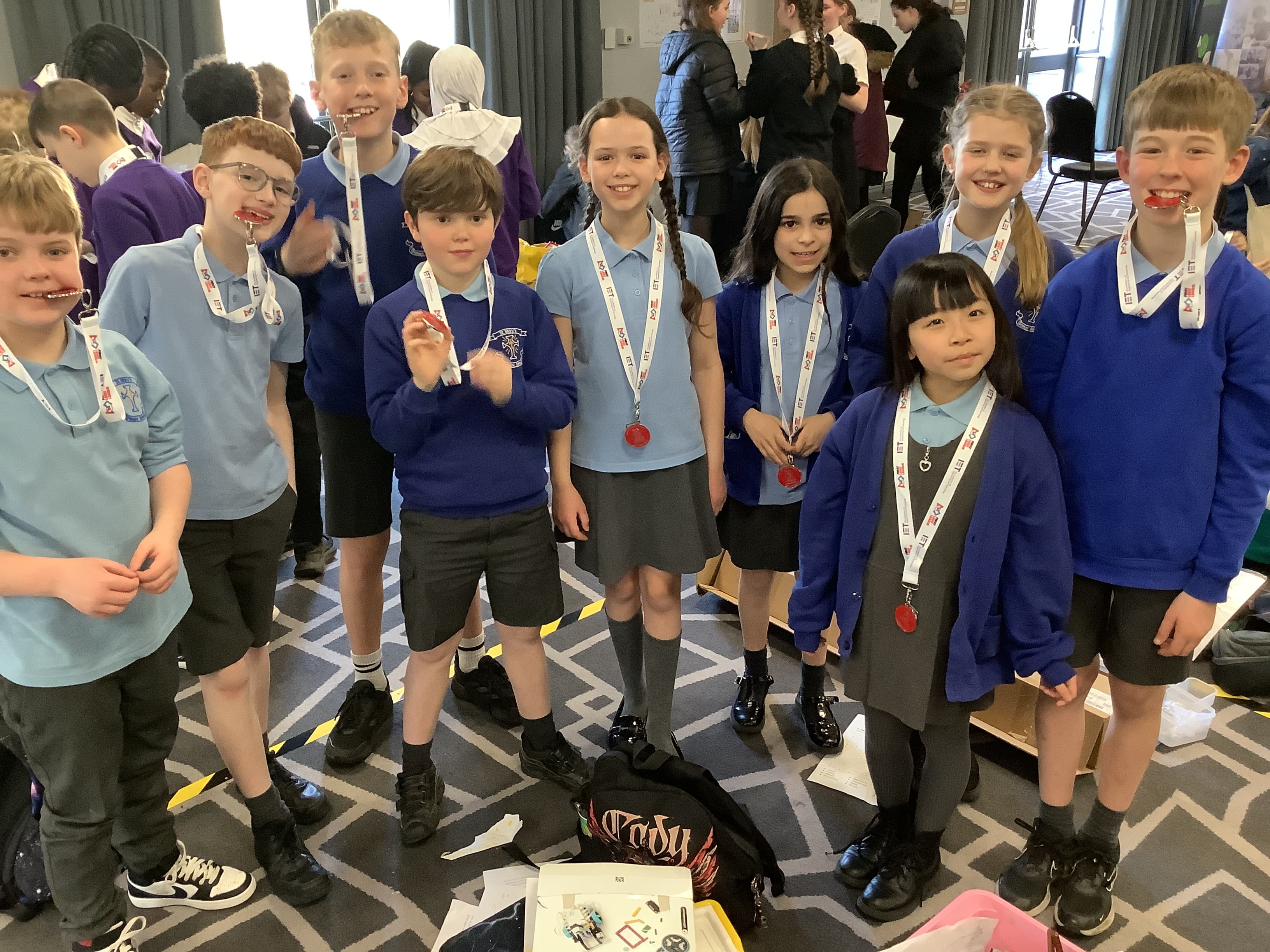 A group of cheerful children wearing school uniforms and lanyards with medals around their necks stand together indoors.