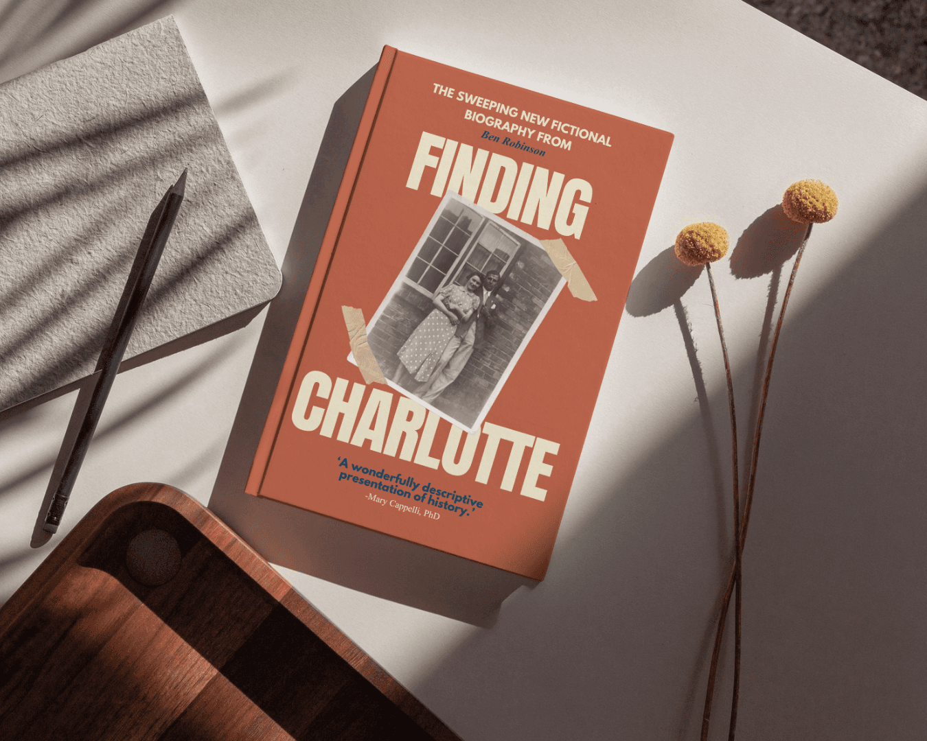 A book titled "Finding Charlotte" with an old photograph of two women on its cover, situated on a white surface with a pencil, a notebook, a wooden tray, and dried yellow flowers.