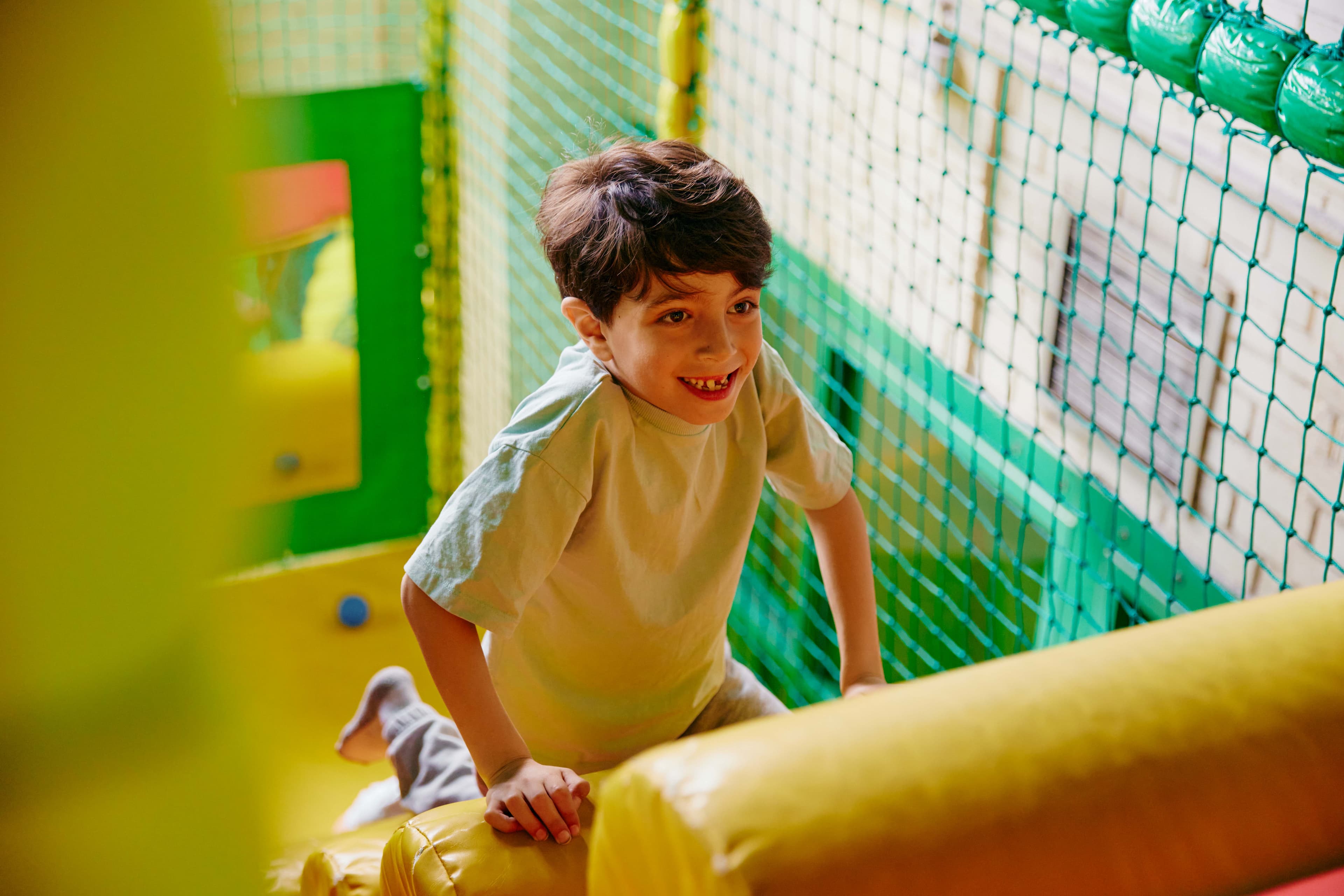 A young boy with dark hair and a light green t-shirt smiles broadly as he plays in a colorful indoor playground with yellow and green padded structures and a green net.
