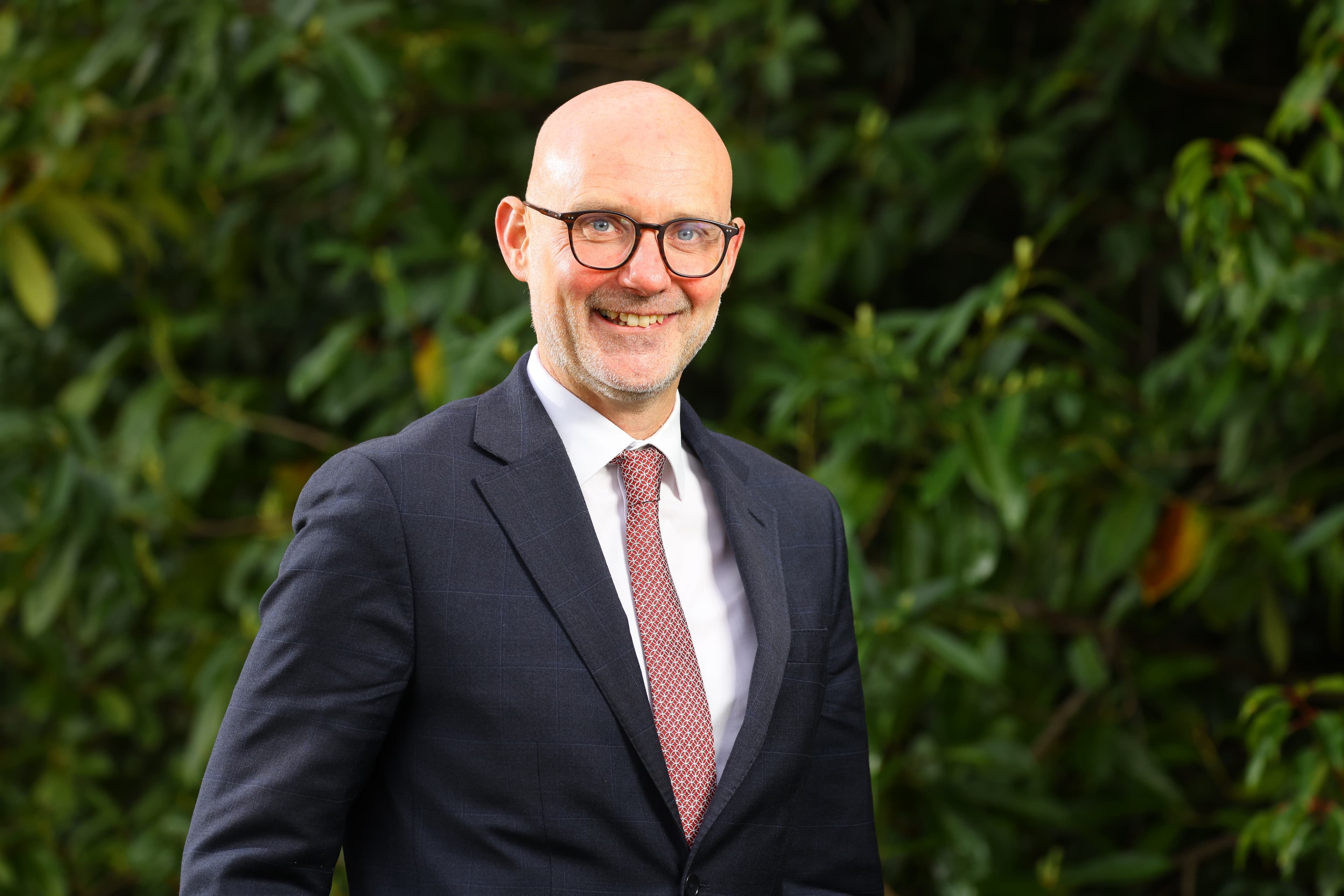 A smiling bald man wearing glasses, a dark suit, and a red patterned tie, stands in front of a leafy green background.