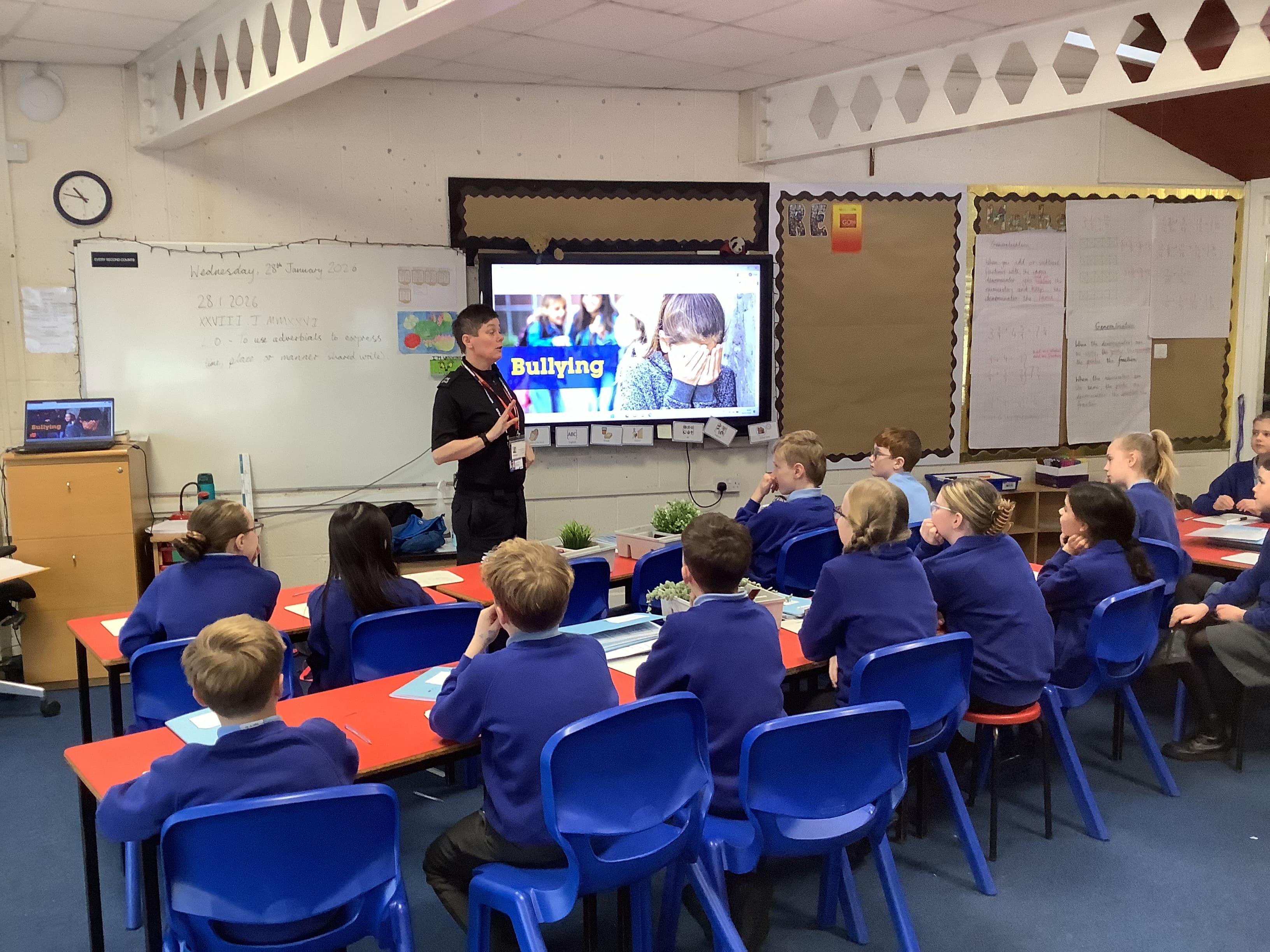 A woman in a dark uniform stands at the front of a classroom, gesturing towards a large screen displaying the word 'Bullying' and an image of a child. Several students, wearing blue uniforms, are seated at desks, attentively watching the presentation.