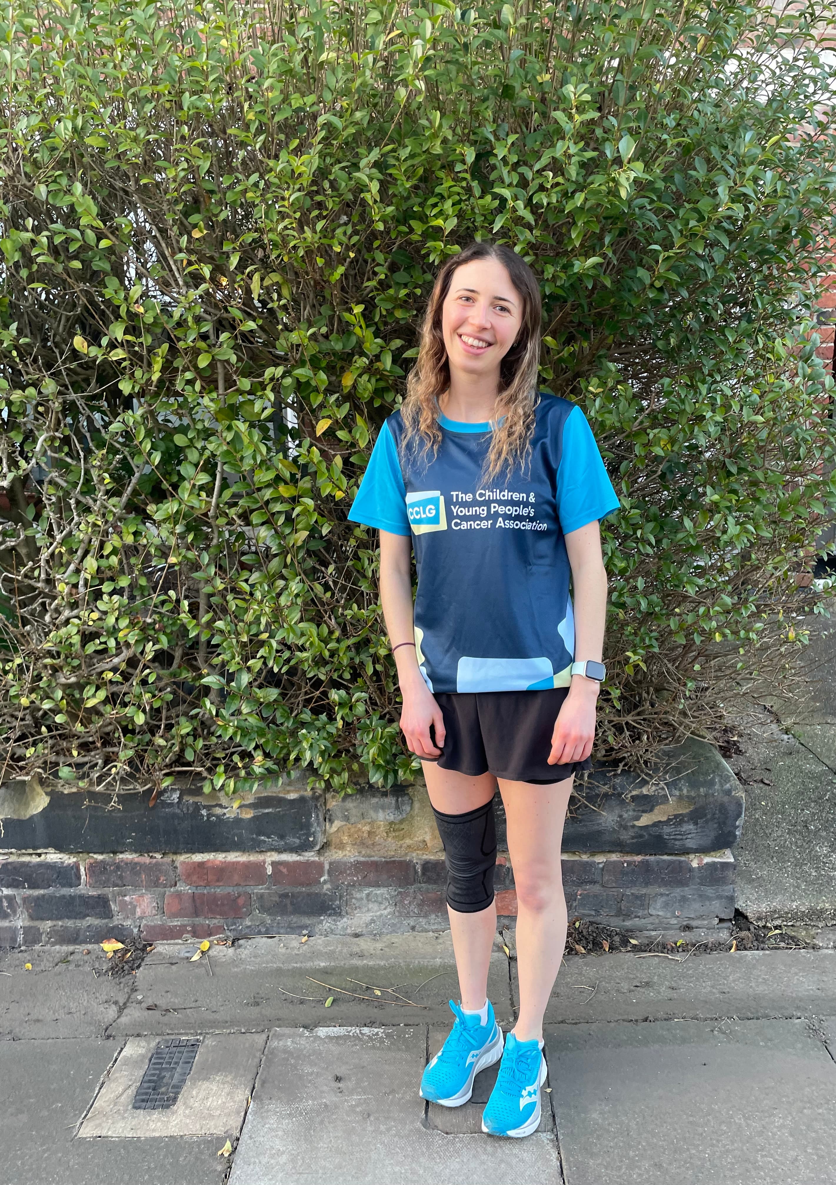 A young woman wearing a blue and black Children & Young People's Cancer Association shirt, black shorts, and bright blue running shoes stands on a sidewalk in front of a green hedge.