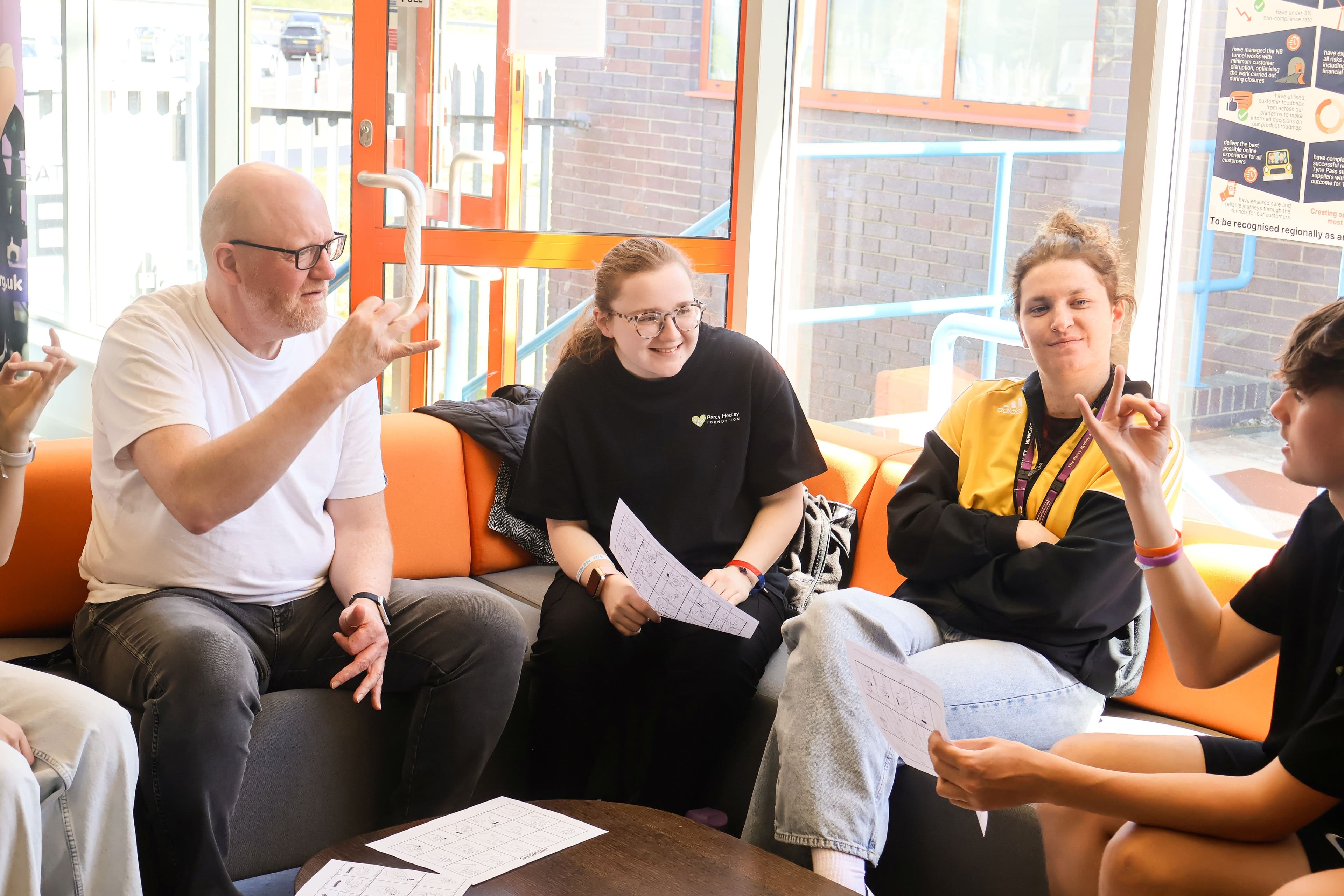 A diverse group of people, including a bald man with glasses, a young woman in a black shirt, and another young woman in a yellow and black jacket, are seated on an orange modular sofa, engaged in conversation and looking at papers spread on a coffee table.
