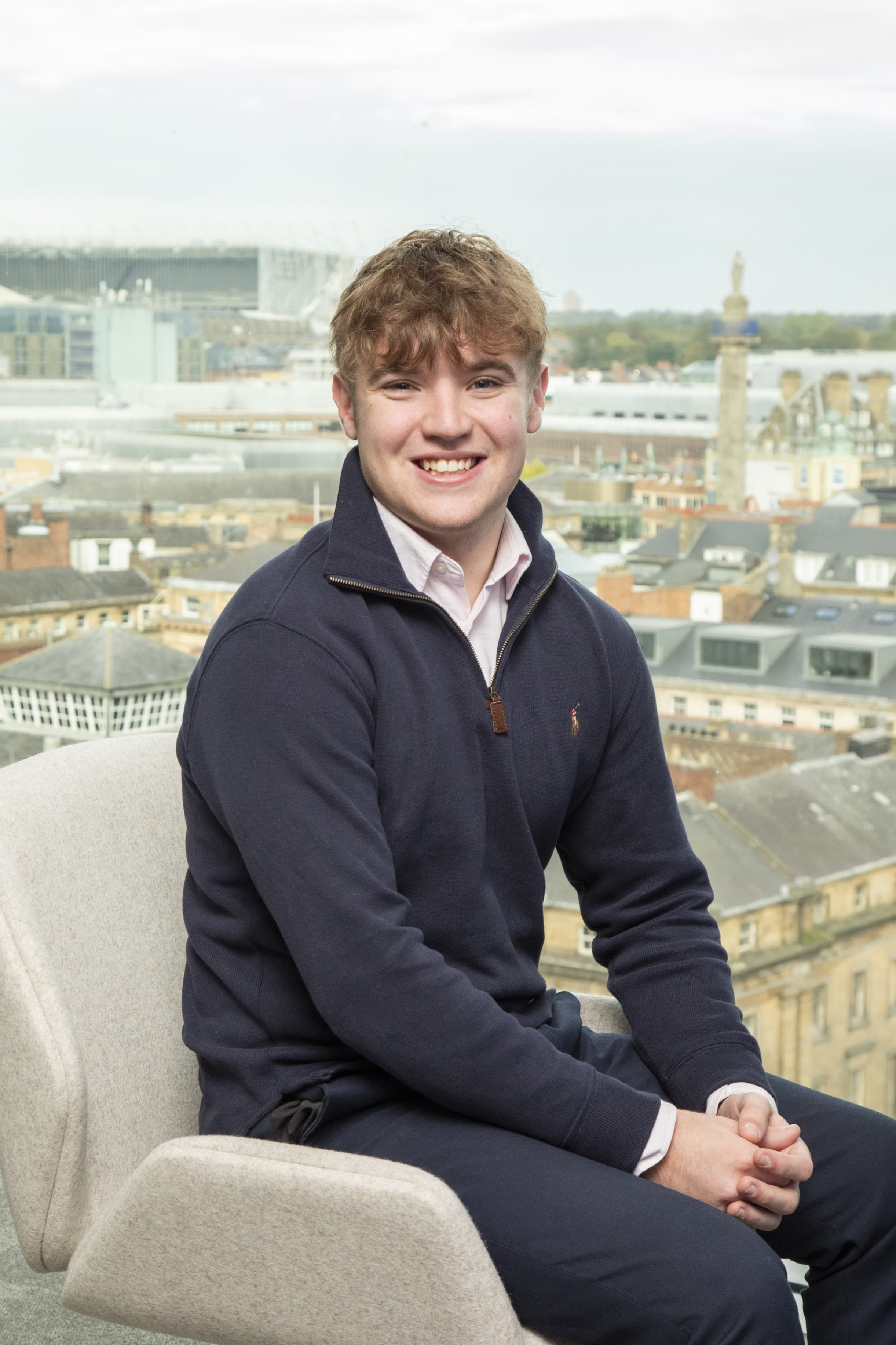 A smiling young man with light brown hair sits on a chair, wearing a navy blue pullover and a light pink shirt, with an urban skyline visible in the background through a window.
