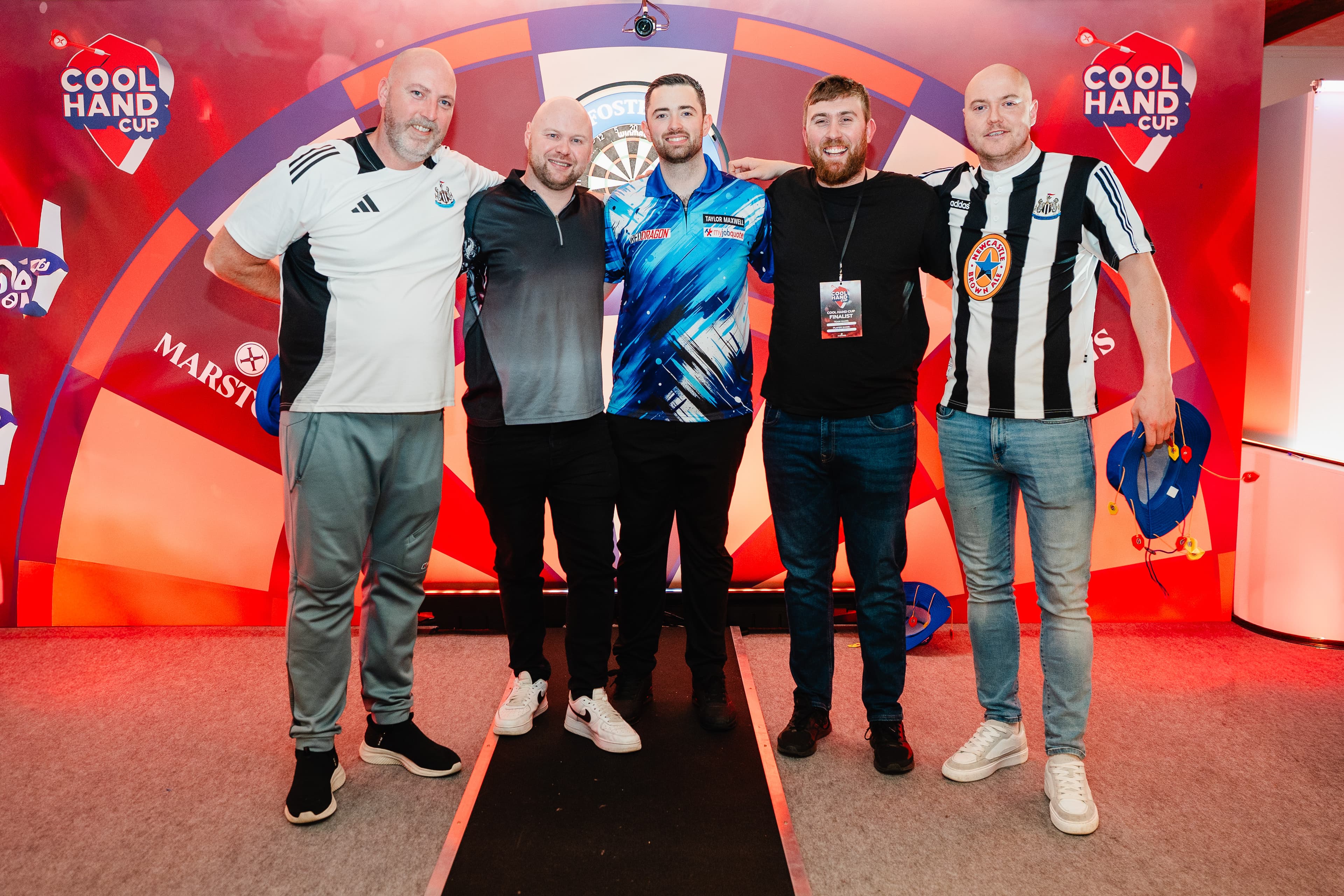A group of five men standing and smiling at the camera in front of a colorful dartboard backdrop with event logos.