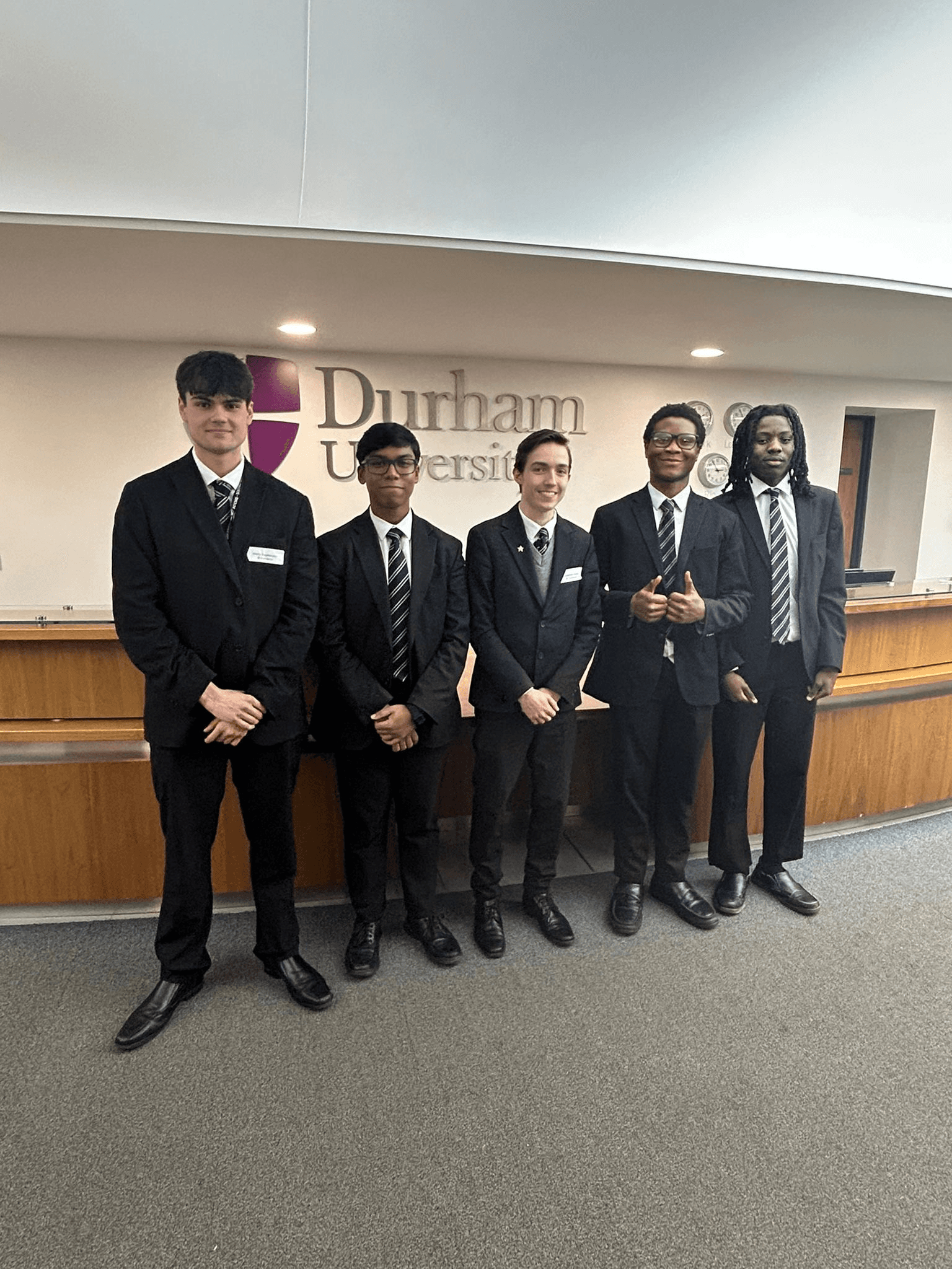 Five young men in black suits and ties stand in front of a white wall with the 'Durham University' logo.