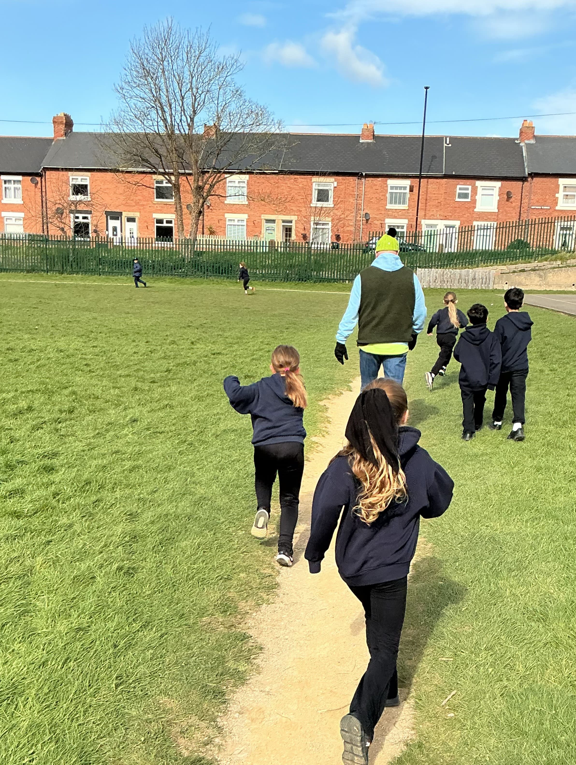 A group of children and an adult from behind walking on a dirt path through a grassy field with brick houses in the background.