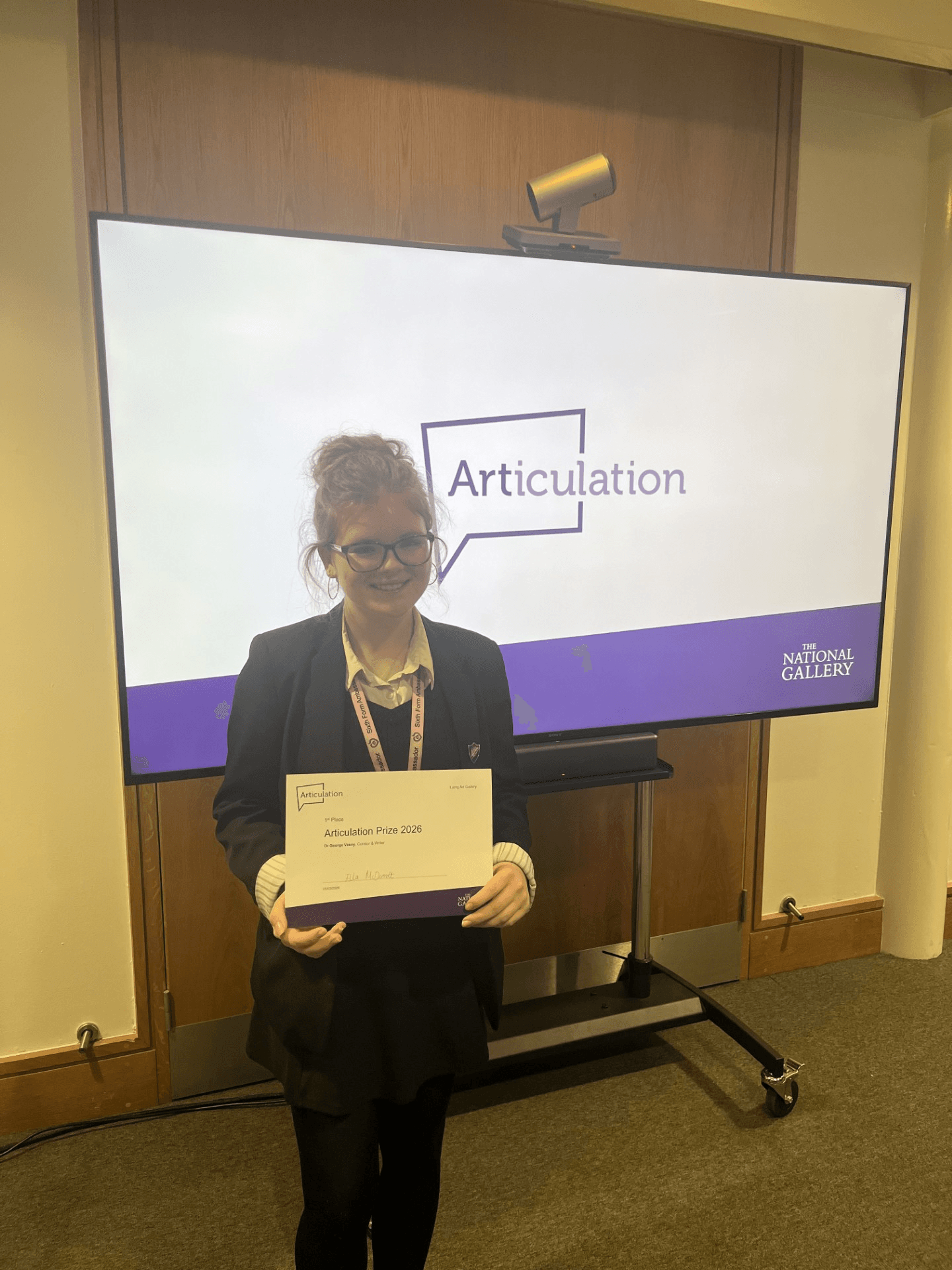 A smiling young woman in glasses and a dark blazer holds a certificate that reads "Articulation Prize 2026" in front of a large screen displaying the word "Articulation" and "The National Gallery" logo.