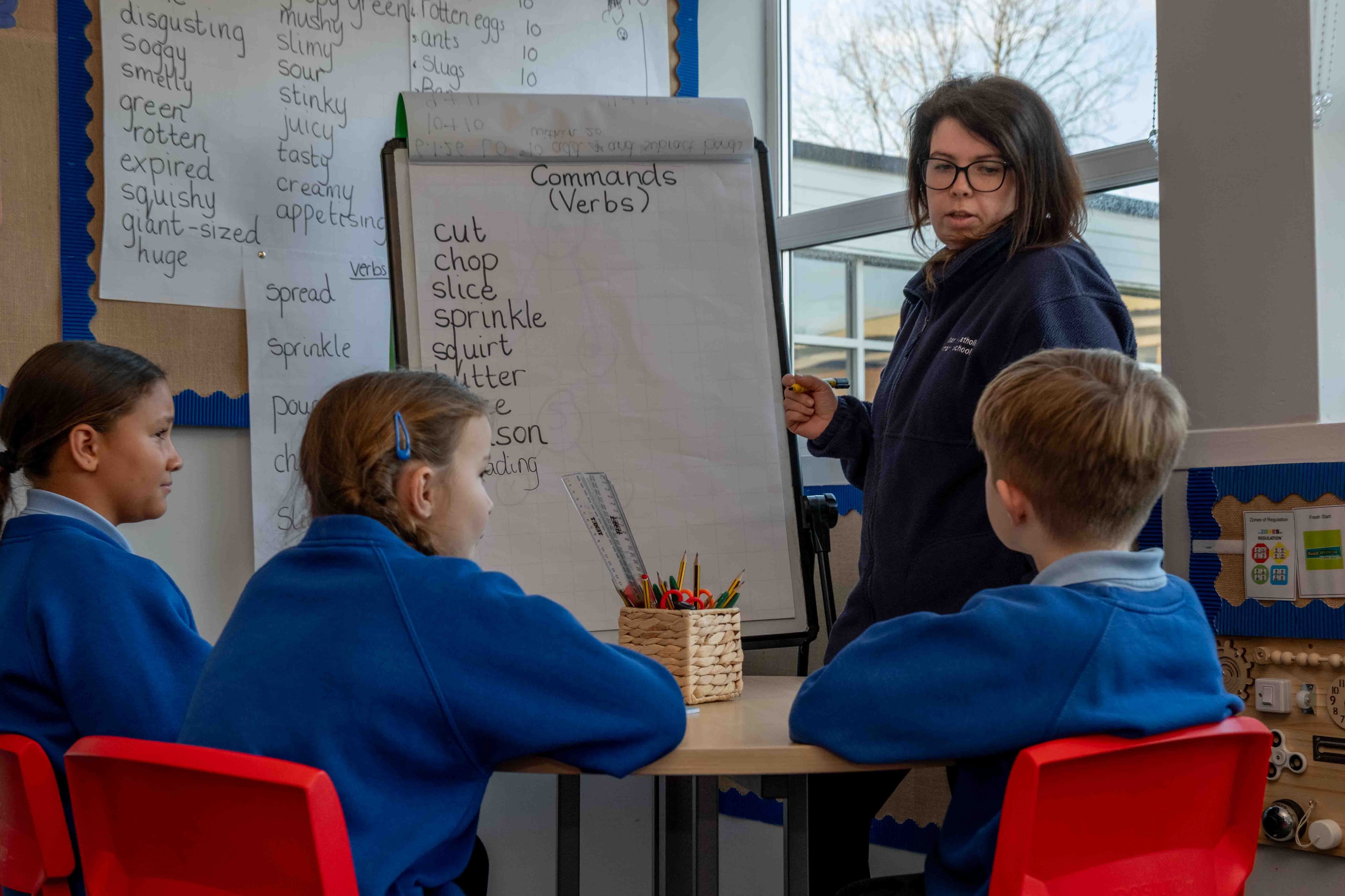 A teacher wearing glasses stands next to a whiteboard, pointing to a list of verbs, while three young students in blue uniforms sit at a table looking at her.