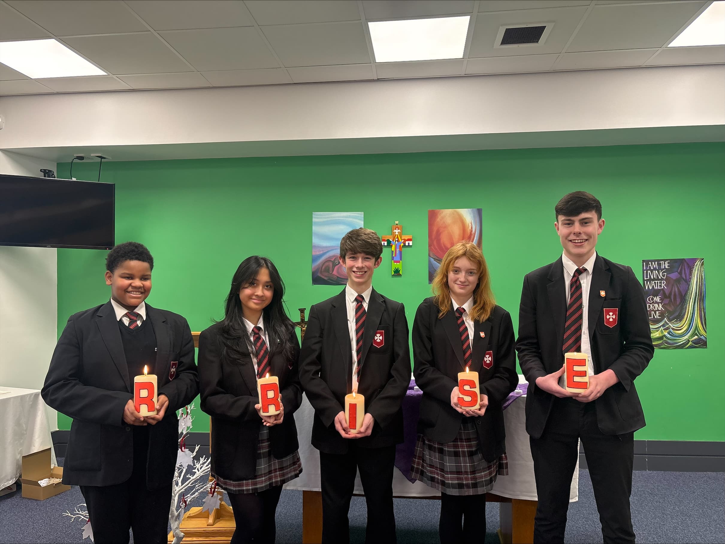 Five students in school uniforms stand in a row holding lit candles, with green walls and religious art in the background.