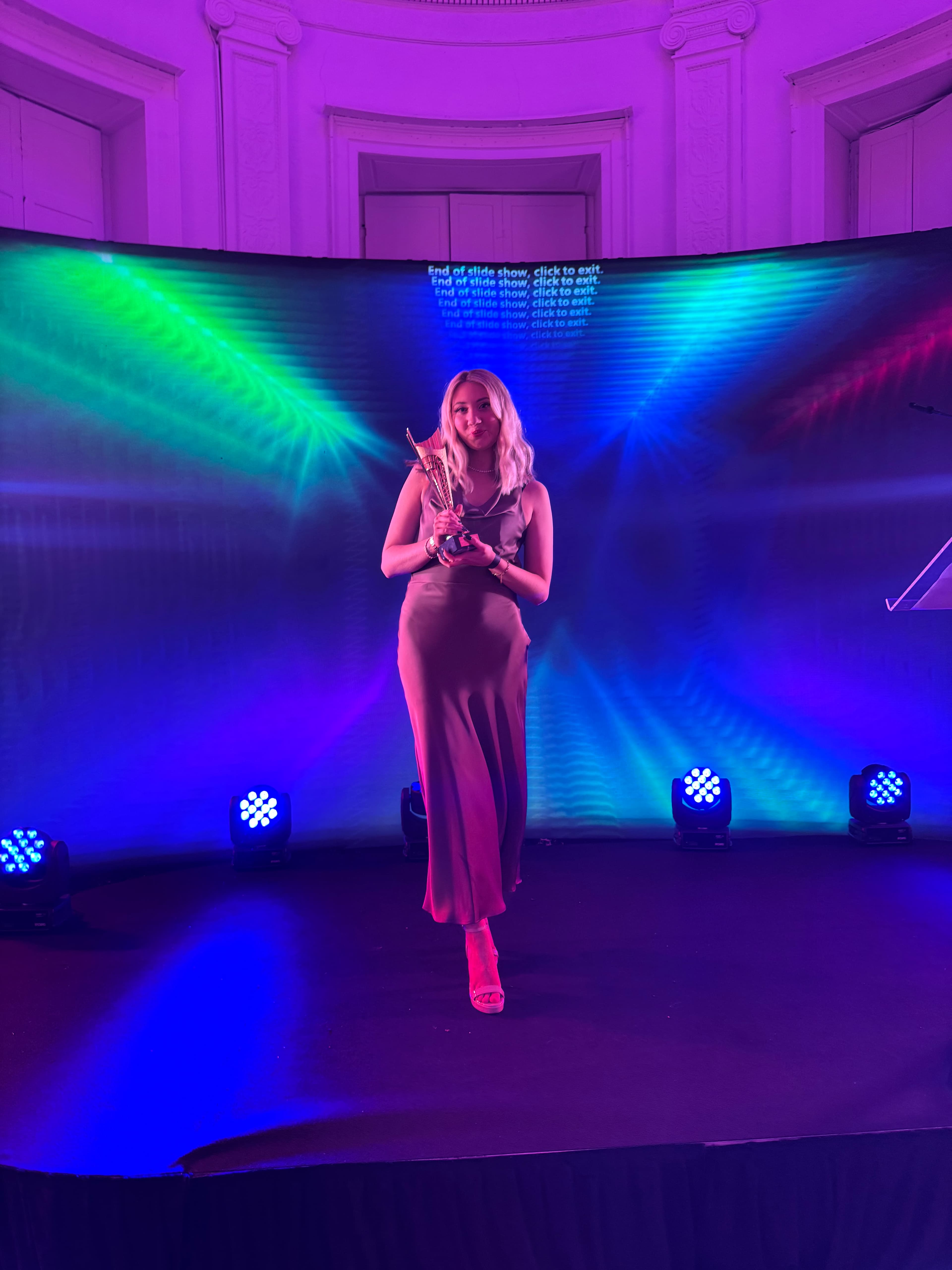 A woman in a long dress stands on a stage holding a golden trophy, bathed in colorful stage lights.