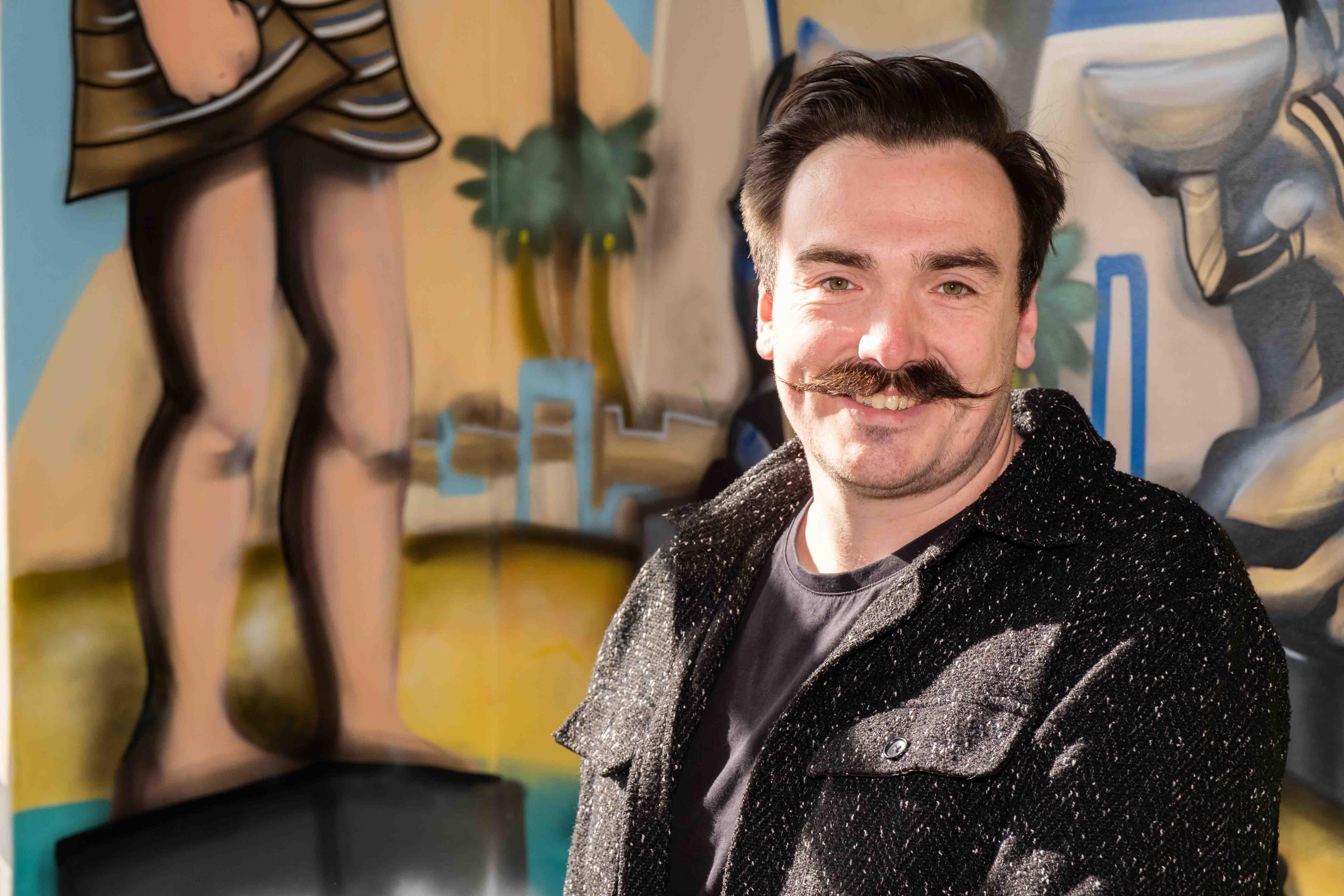 A smiling man with a large curly mustache and dark hair, wearing a dark tweed jacket, stands in front of a colorful, abstract mural.