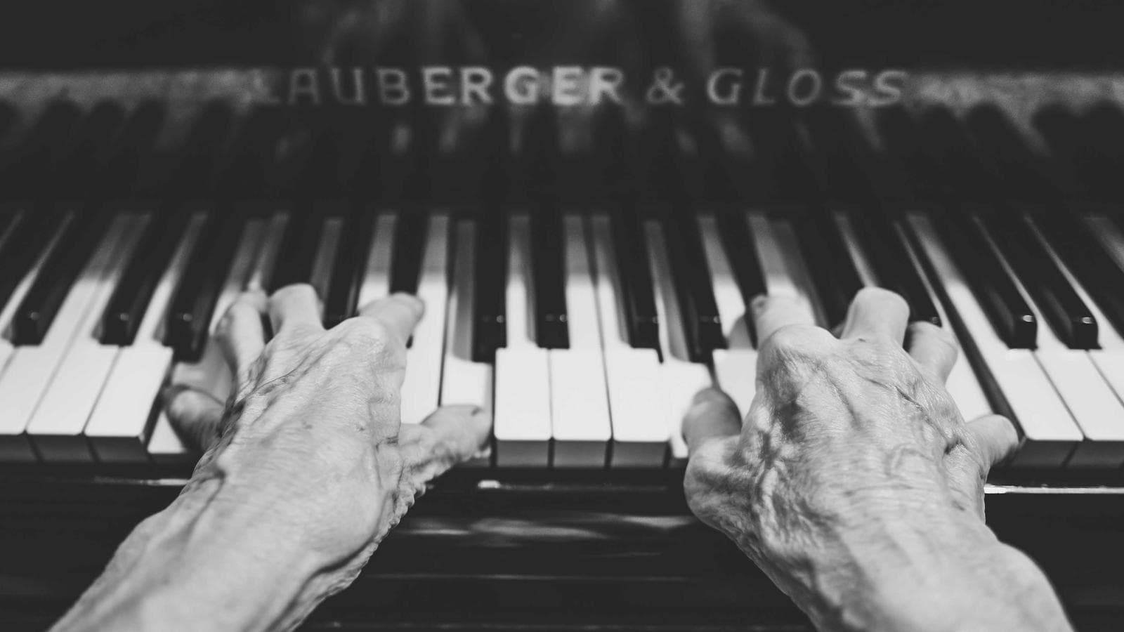 A pair of aged, wrinkled hands play a piano, pressing down on several white and black keys with the "RAUBERGER & GLOSS" brand name visible on the piano's nameboard.