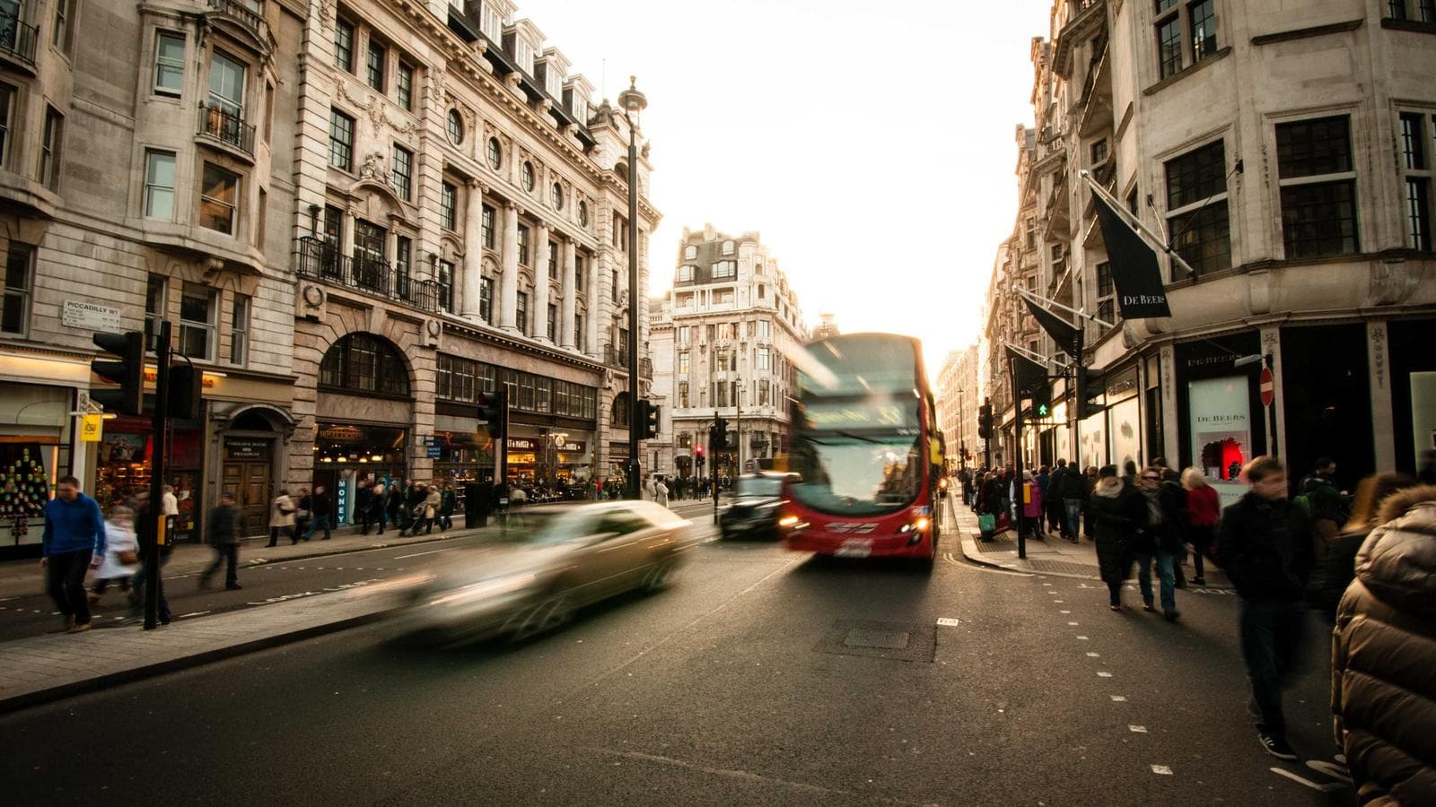 A busy city street scene with a red double-decker bus and a motion-blurred car in the foreground, surrounded by historic buildings and pedestrians.