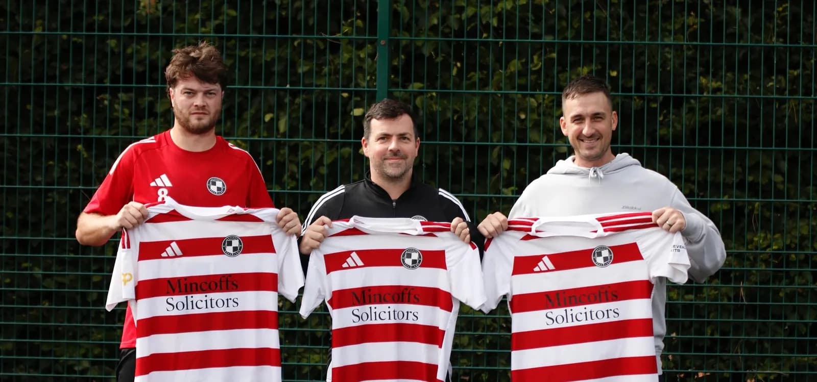 Three men stand side-by-side in front of a green fence, each holding up a white and red striped soccer jersey.