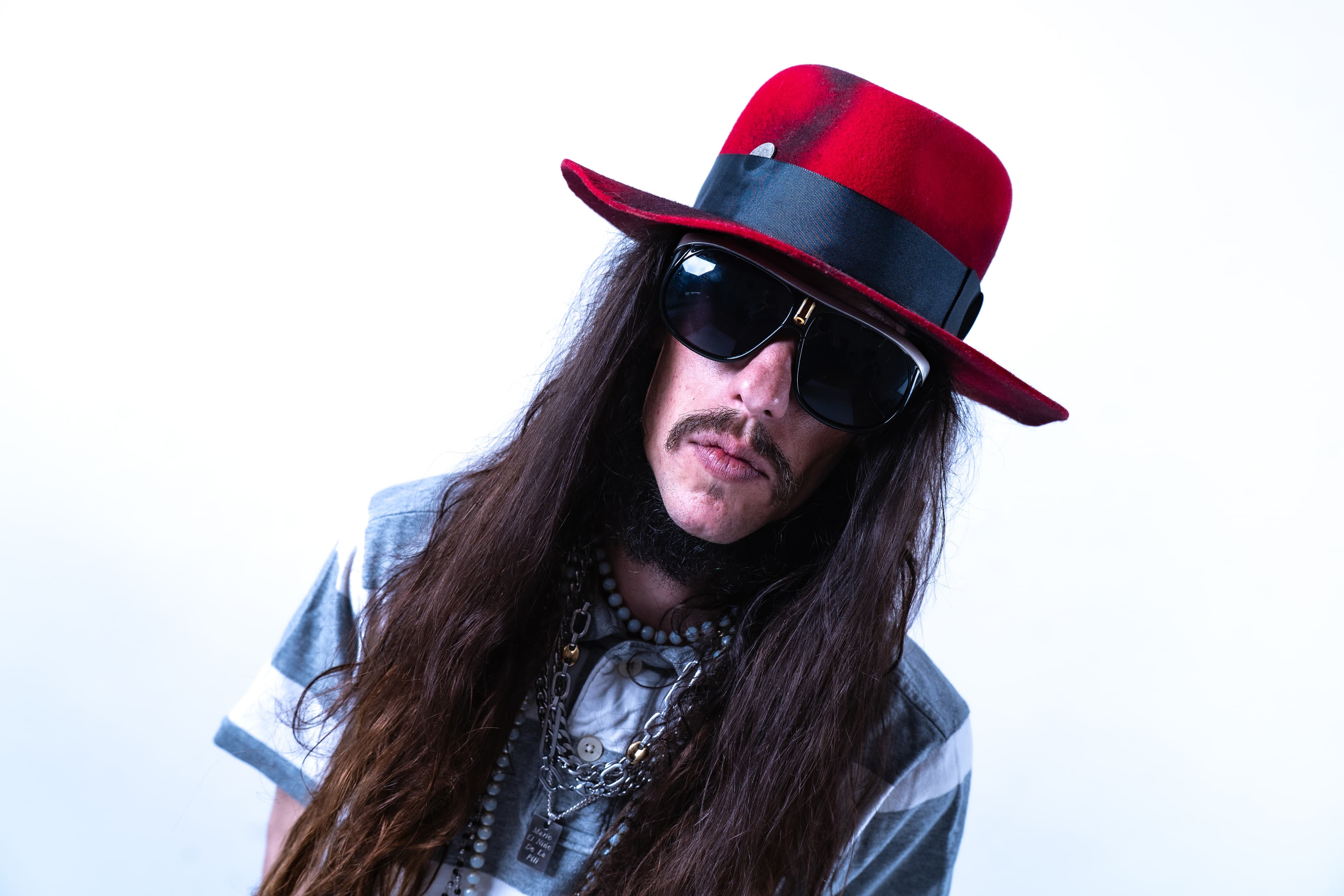 A person with long brown hair, a mustache, sunglasses, and a red fedora with a black band, wearing a grey striped shirt and multiple necklaces, looking directly ahead.