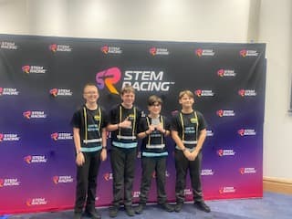 Four boys in black STEM Racing shirts with lanyards stand in front of a blue and black STEM Racing banner.