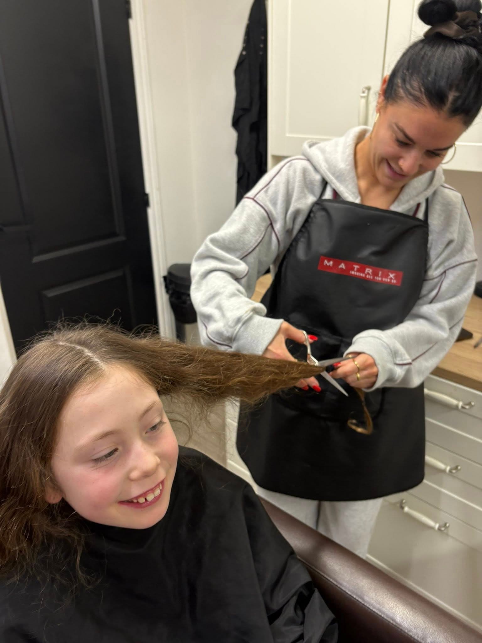 A young girl with long brown hair smiles widely as a woman with a black apron and gray hoodie cuts a thick section of her hair with scissors.
