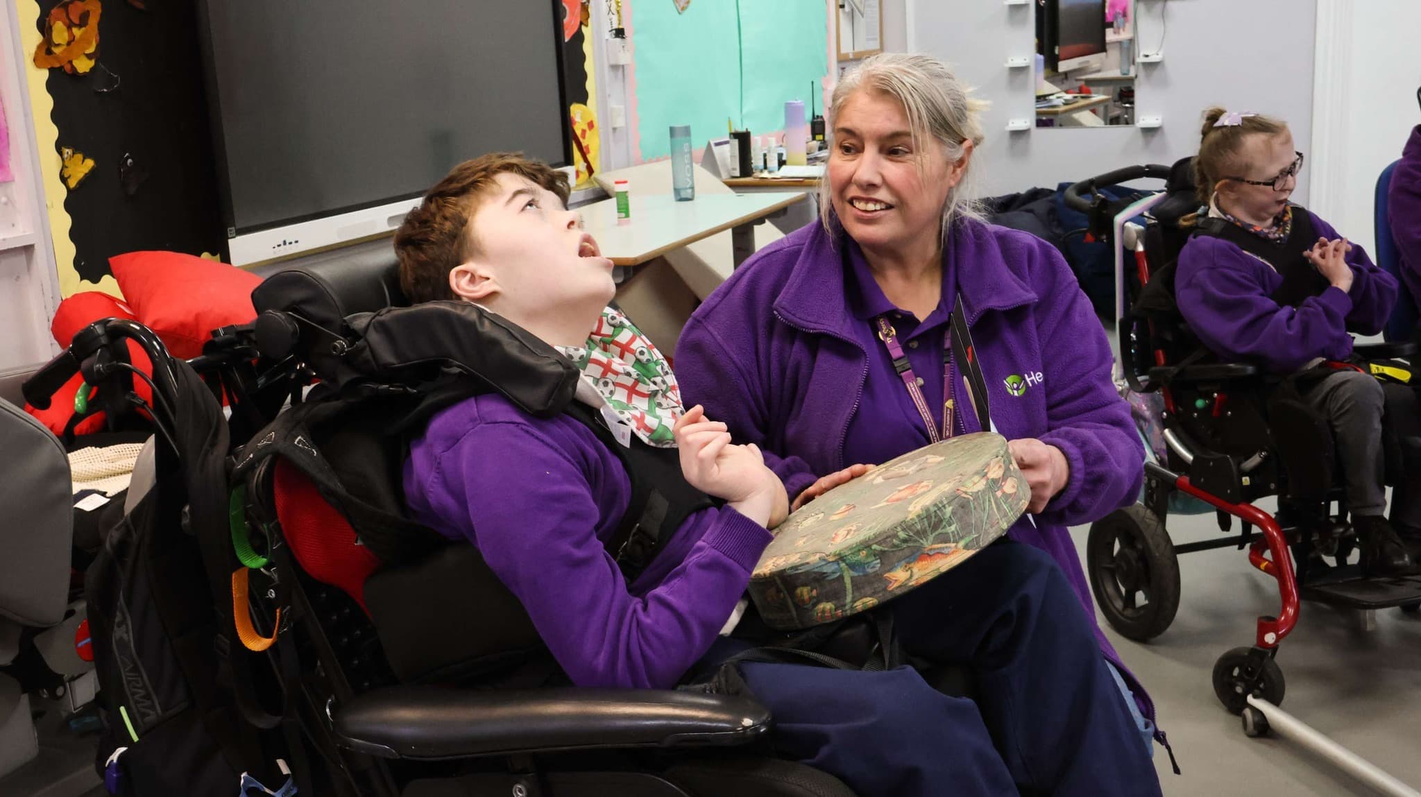 A young person in a wheelchair with their head tilted back and mouth open is enjoying an activity with an adult caregiver in purple, who smiles at them while holding a shallow drum in a classroom setting.