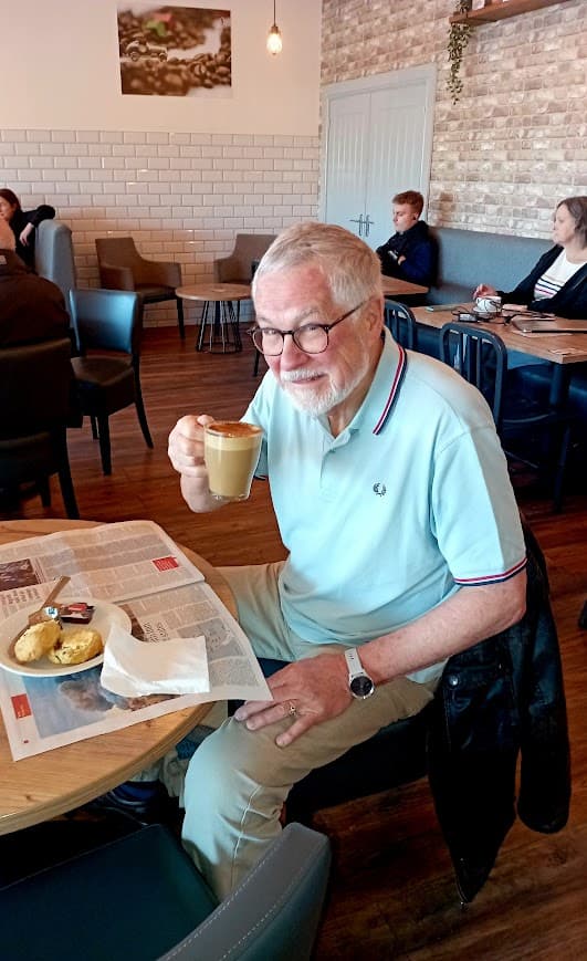 A man with glasses and a light blue polo shirt sits at a table in a cafe, smiling while holding a glass of coffee, with a newspaper and scones on the table.