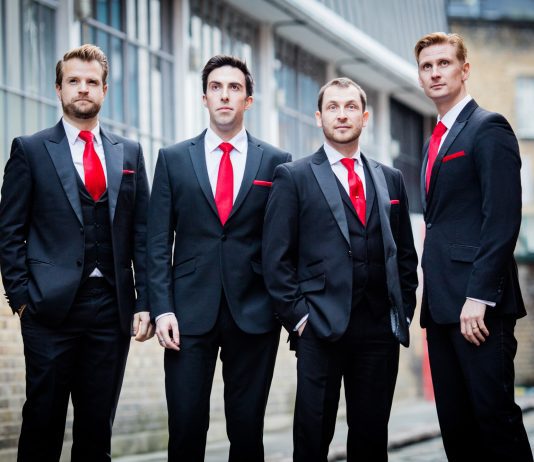 Four men in smart suits with red ties and pocket squares. They are supposed to all be looking at something in sky for this shot, but one of them is looking at the camera.