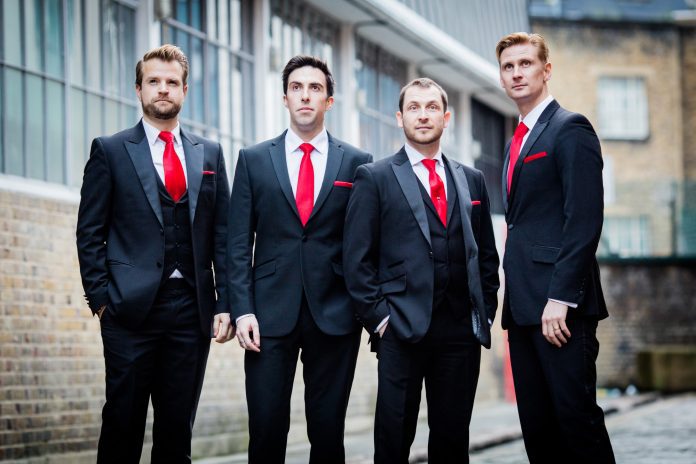 Four men in smart suits with red ties and pocket squares. They are supposed to all be looking at something in sky for this shot, but one of them is looking at the camera.